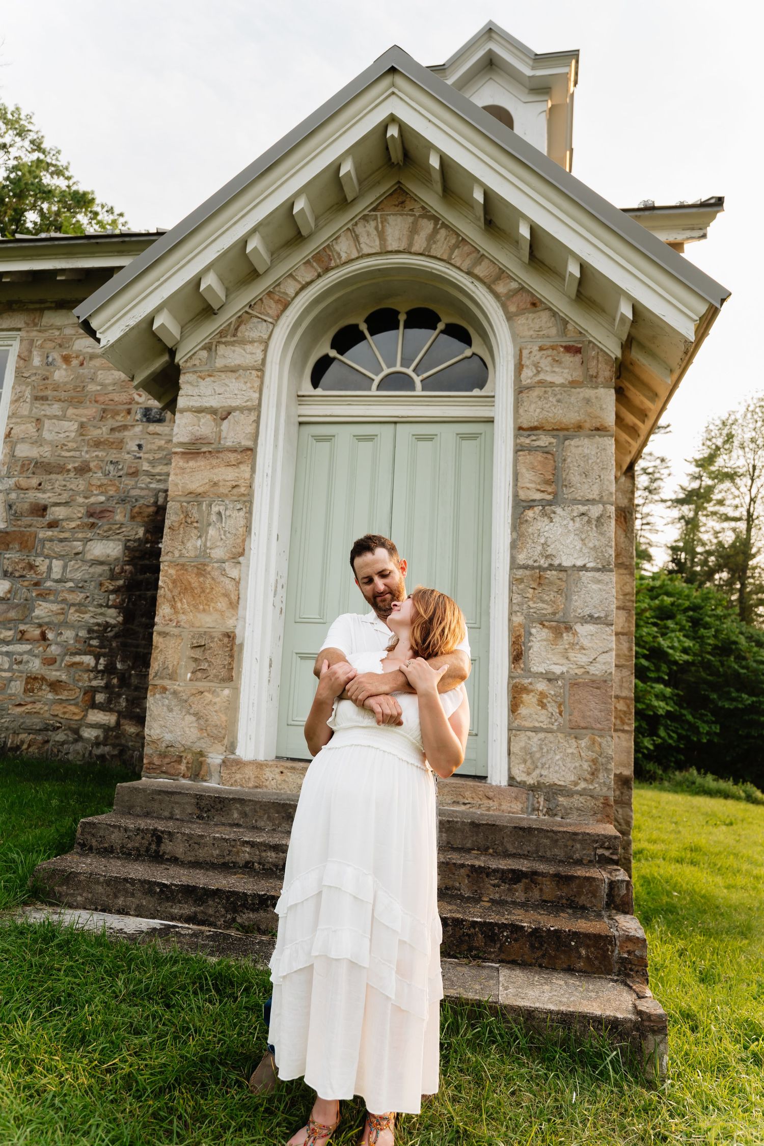 A couple stands on the stone steps of a historic chapel with arched doorway and rustic stone exterior in golden sunlight.