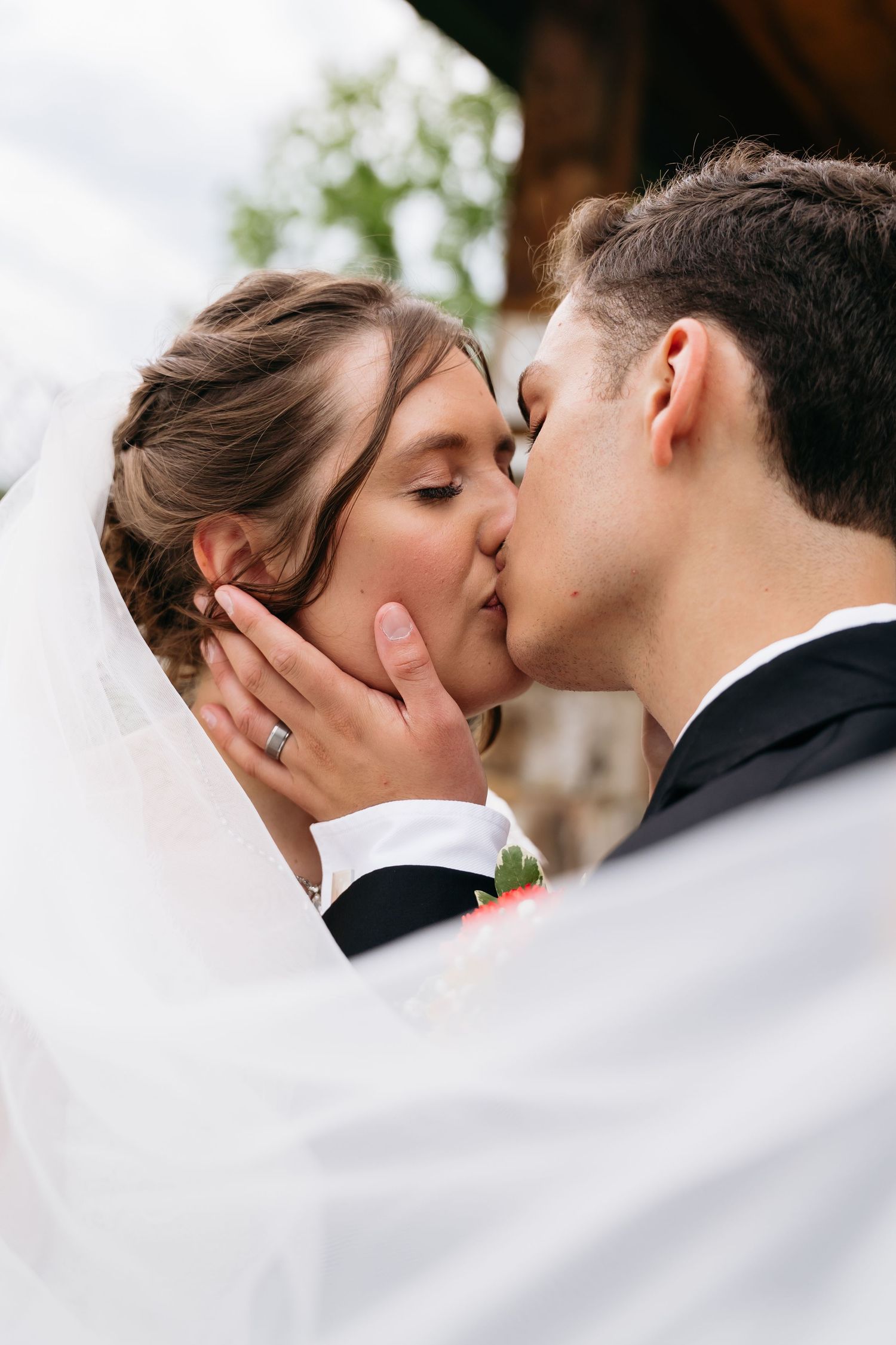 A romantic wedding day kiss between newlyweds outdoors with the bride's flowing white veil in the foreground.