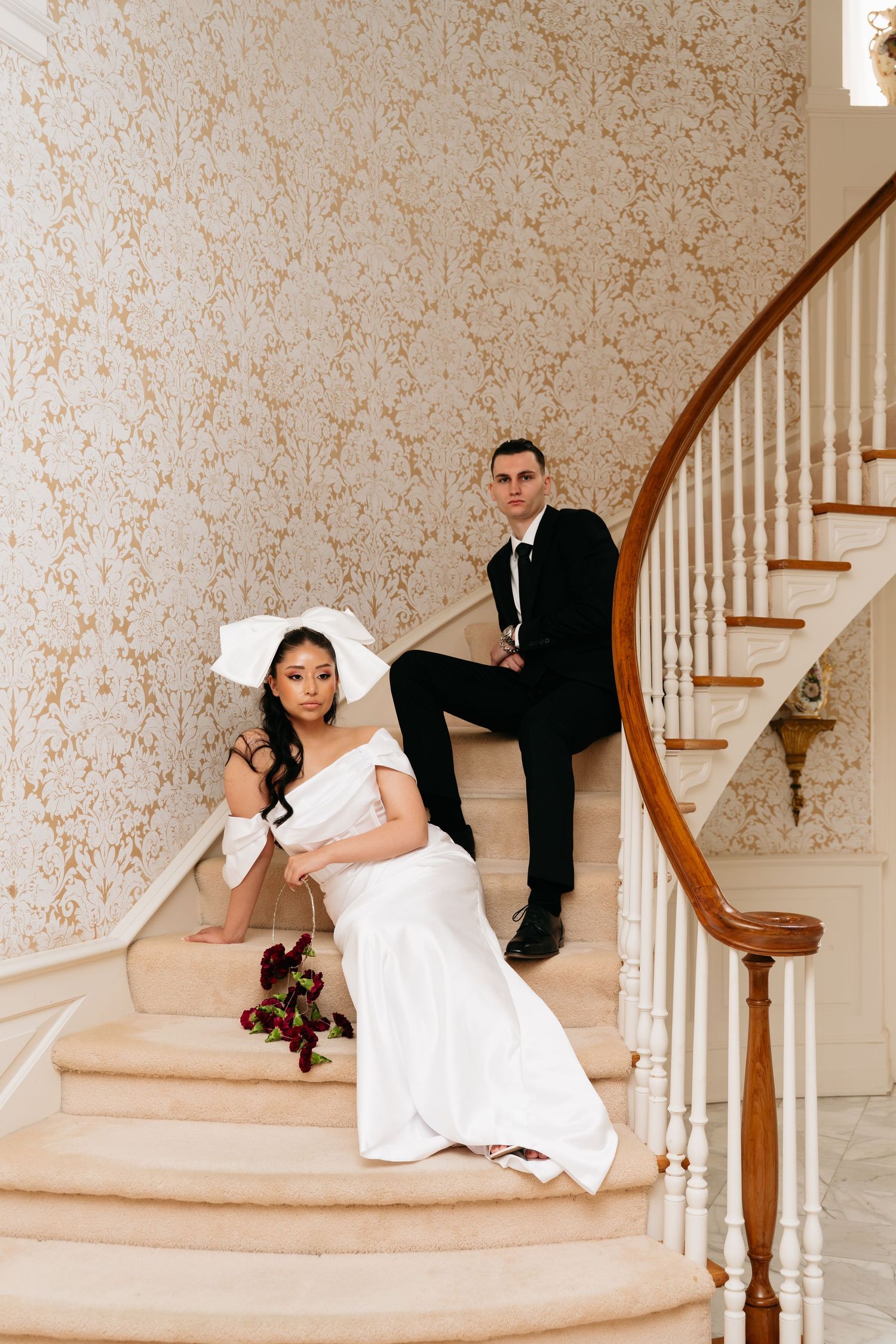 Bride in white gown and hat sits on elegant staircase with groom in black suit at luxurious wedding venue.