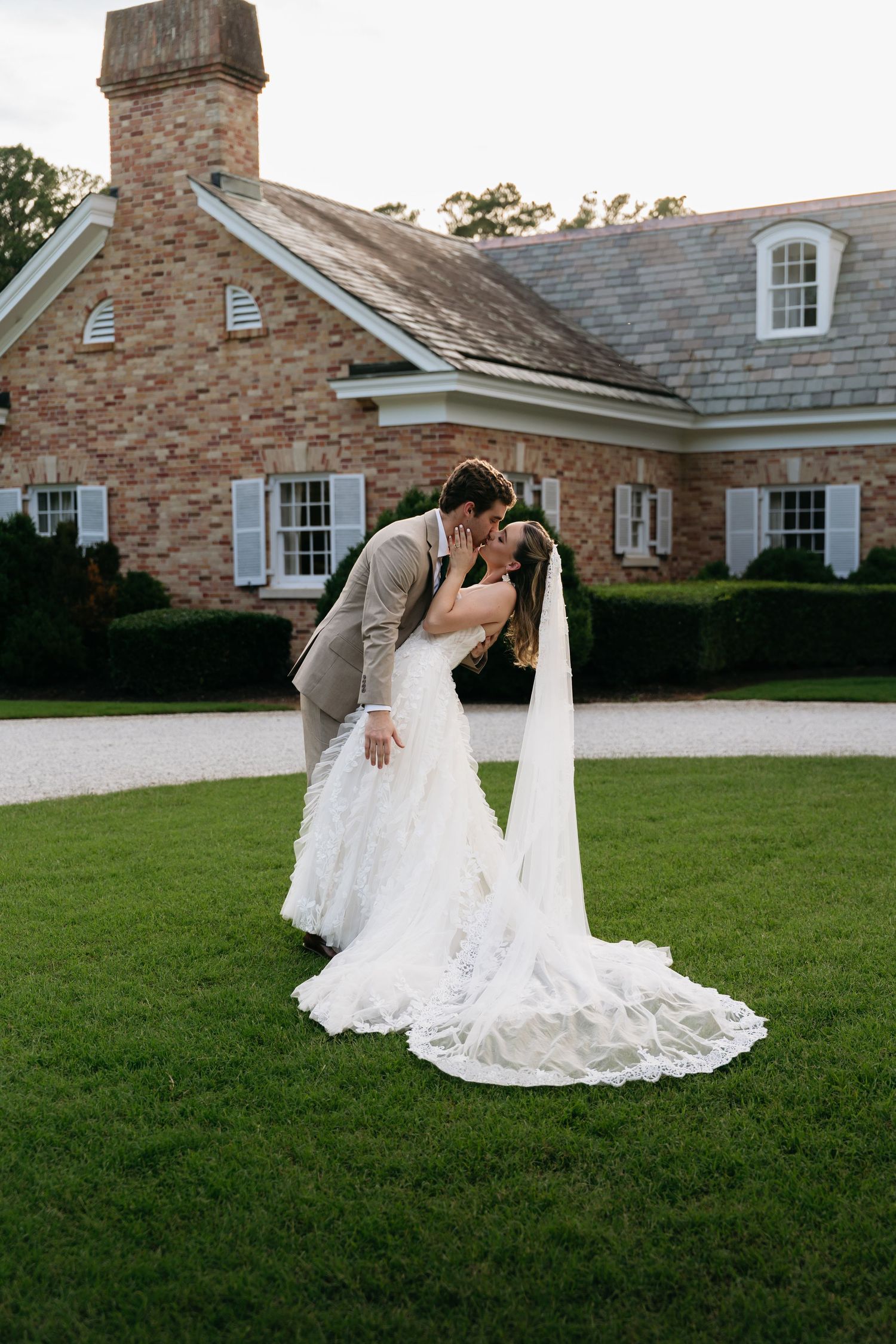 Bride and groom share a romantic dip kiss on green lawn outside brick estate venue with long lace veil.
