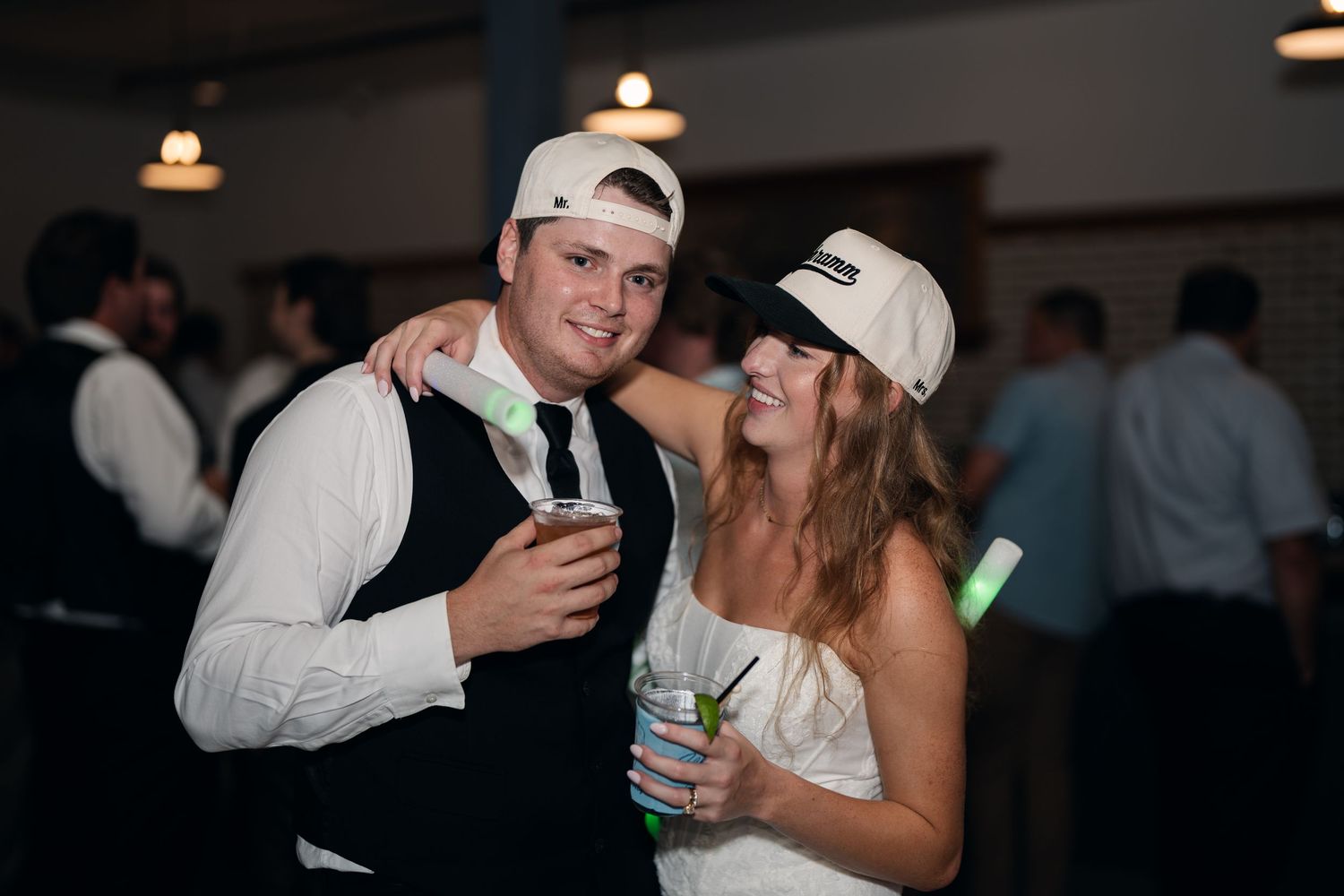 Couple wearing white baseball caps and holding drinks celebrating at lively indoor wedding reception party.