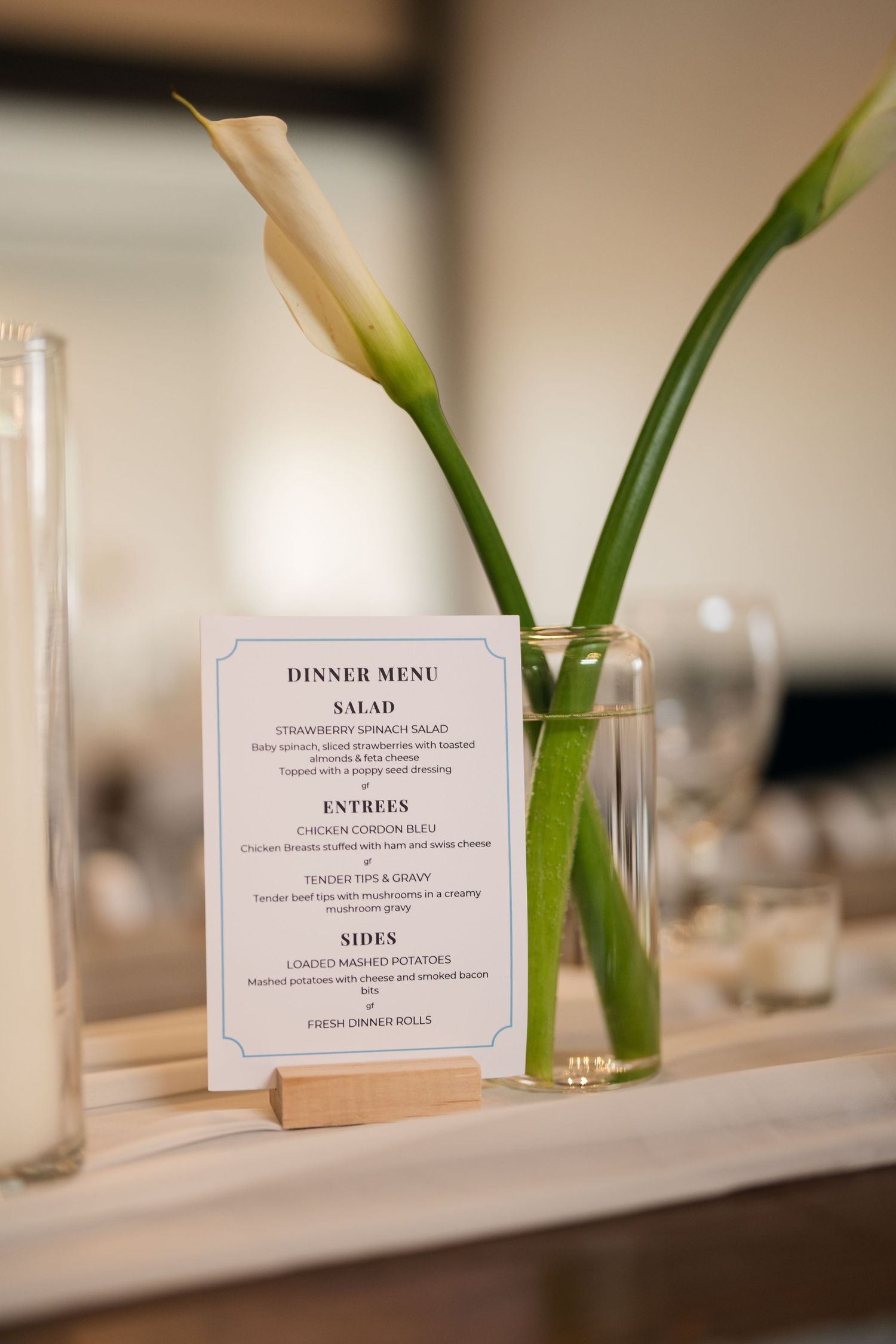 Elegant wedding dinner menu card displayed beside calla lily stems in a glass vase on a reception table.