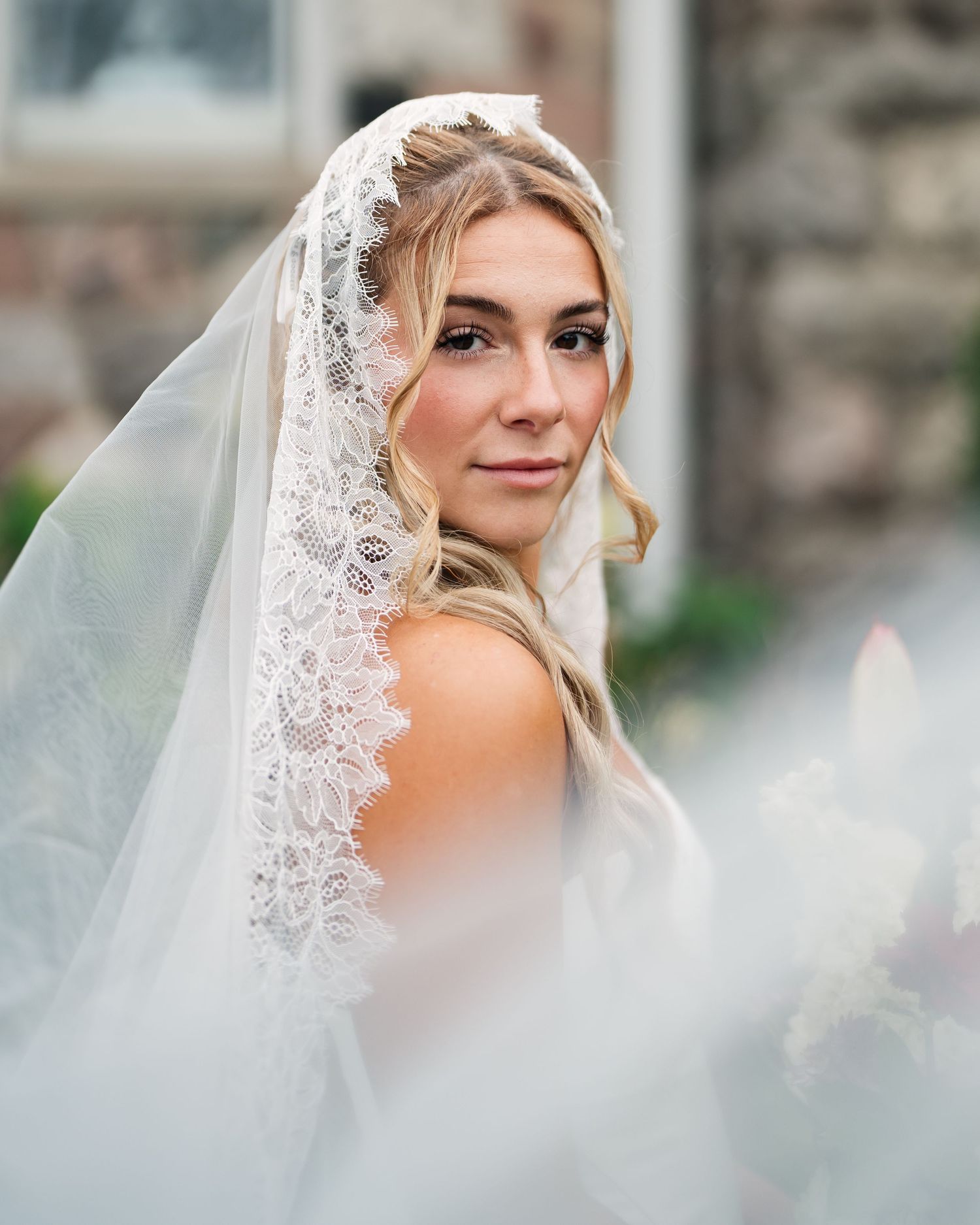 A bride wearing a lace-trimmed veil and strapless gown poses elegantly outdoors near a stone building.