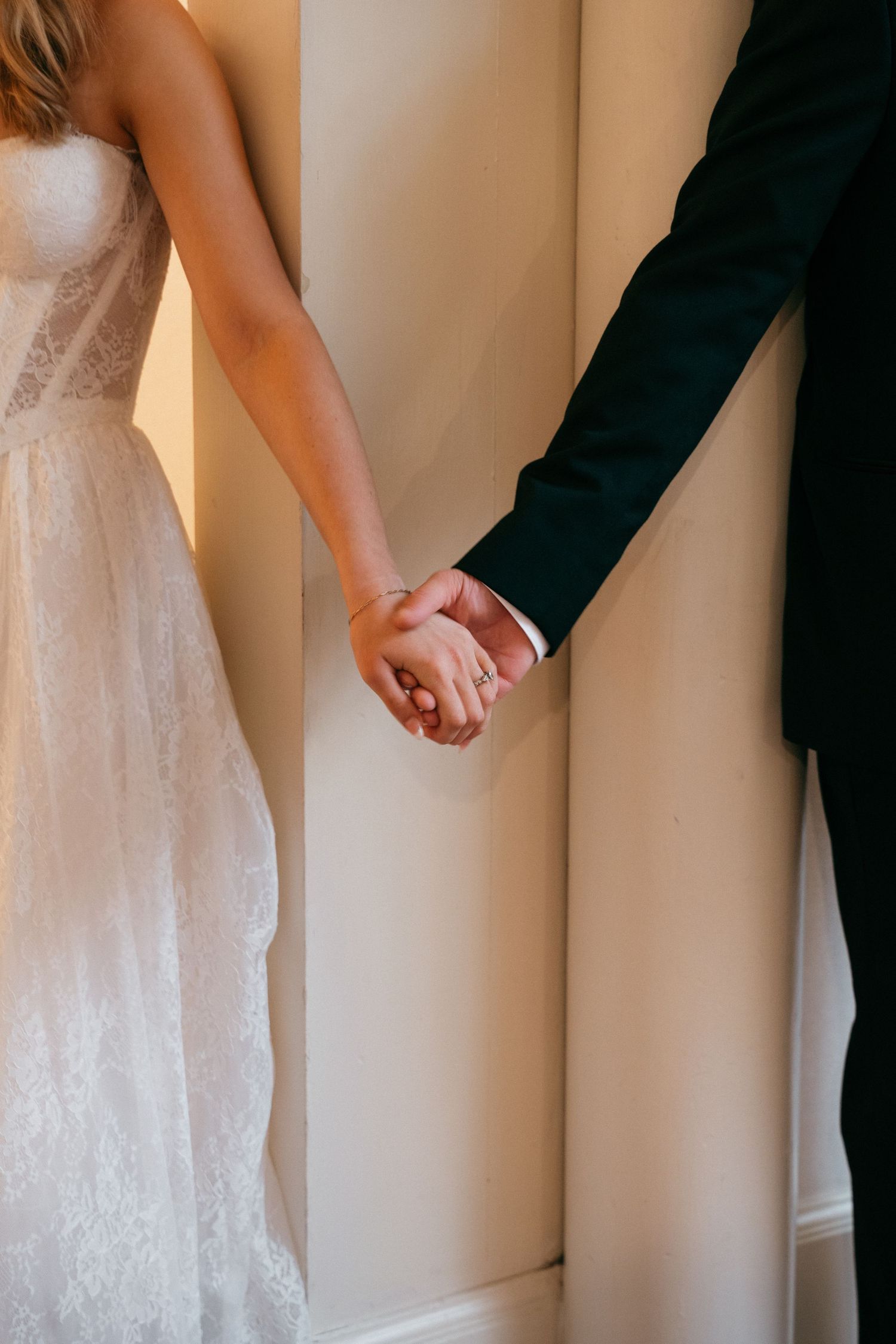 Bride and groom holding hands in wedding attire, showcasing a diamond ring and elegant lace dress.