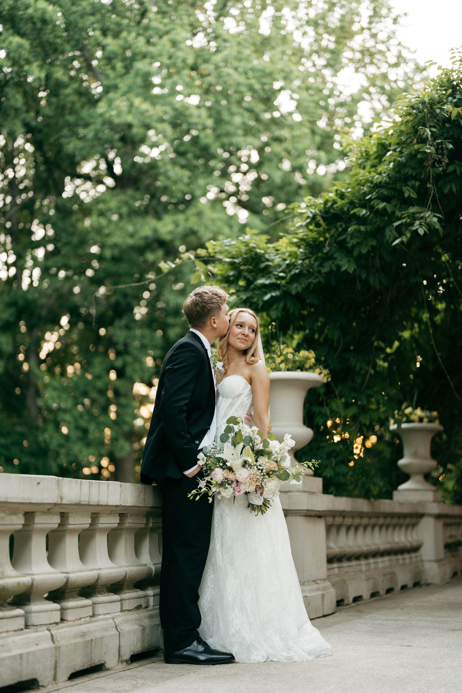 Groom kissing bride on cheek as she holds lush bouquet beside stone balustrade in lush garden.
