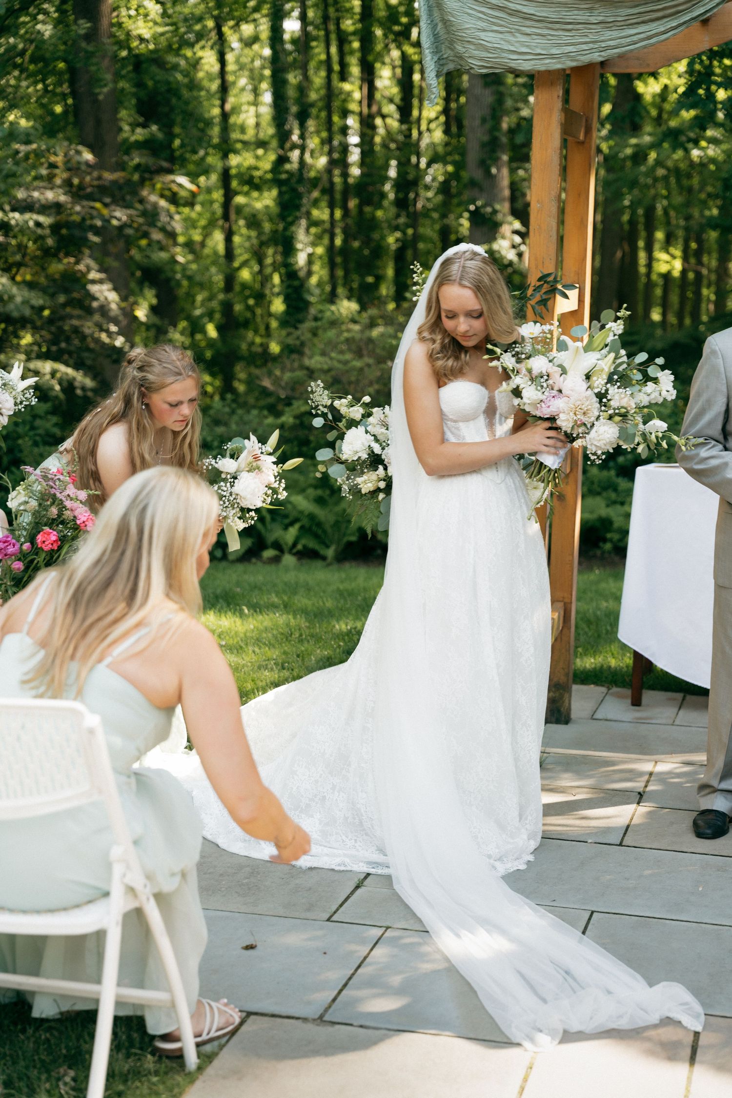 Bridesmaids in white dresses adjust bride's lace train as she holds floral bouquet at outdoor garden ceremony.