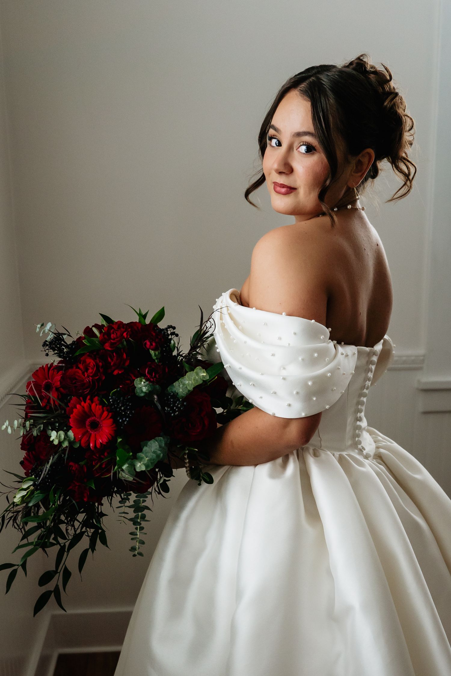 A bride in an elegant white ballgown poses with a dramatic deep red and green wedding bouquet against a neutral backdrop.
