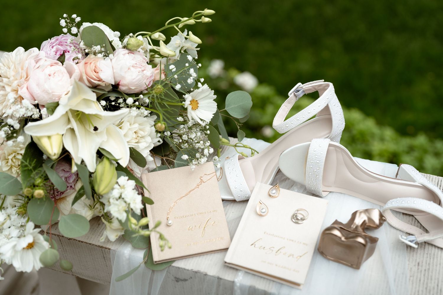 Elegant wedding flat lay with bridal bouquet, white heels, pearl earrings, and invitation cards on white surface.