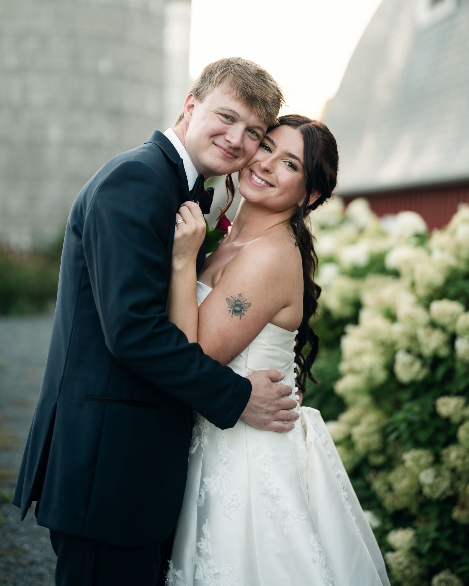 A smiling bride and groom pose together outdoors near white hydrangeas, the bride in a strapless gown and groom in a dark suit.