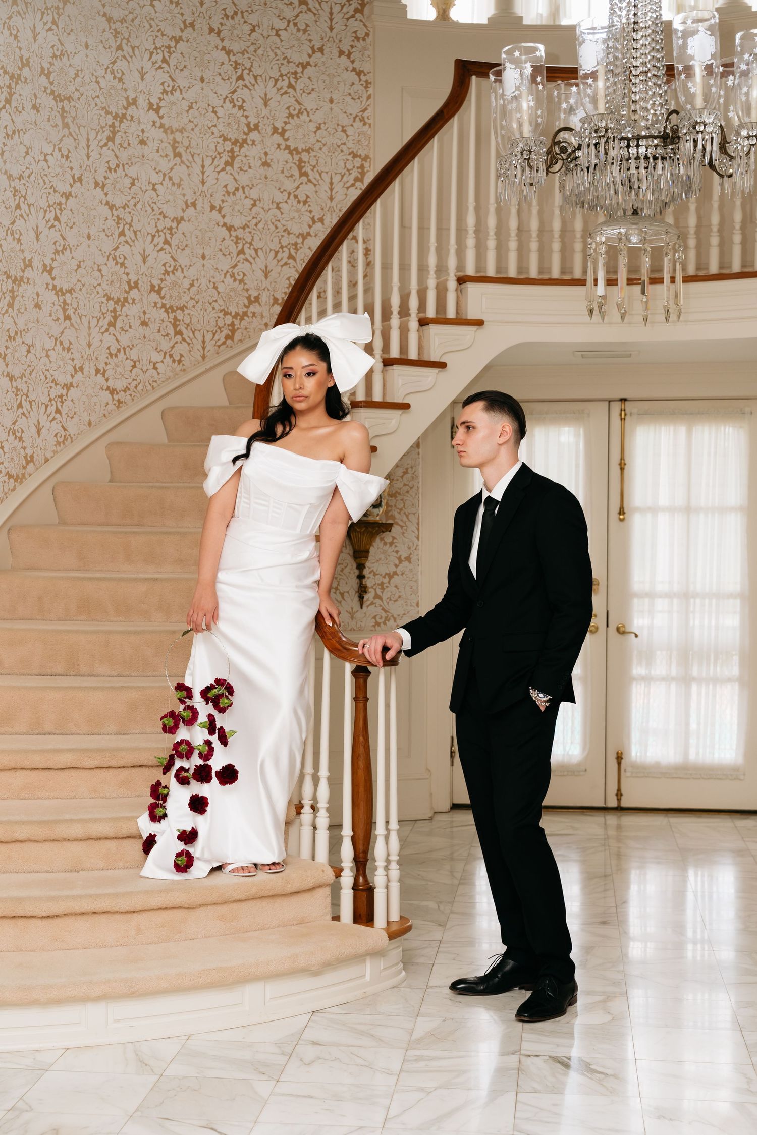 Bride in white off-shoulder gown with bow and groom in black suit holding hands on elegant curved staircase.