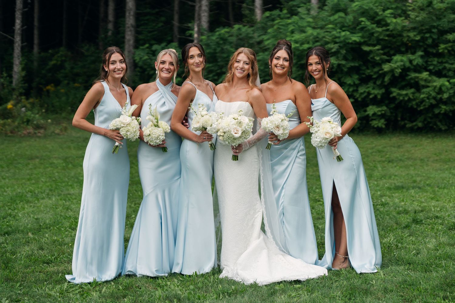 Bride in white gown with five bridesmaids in light blue dresses holding white bouquets outdoors.