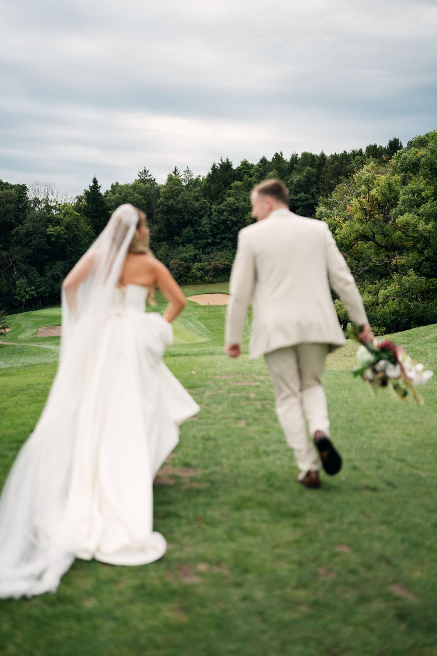 Bride and groom walk across a lush green golf course landscape under a cloudy sky, bouquet in hand.