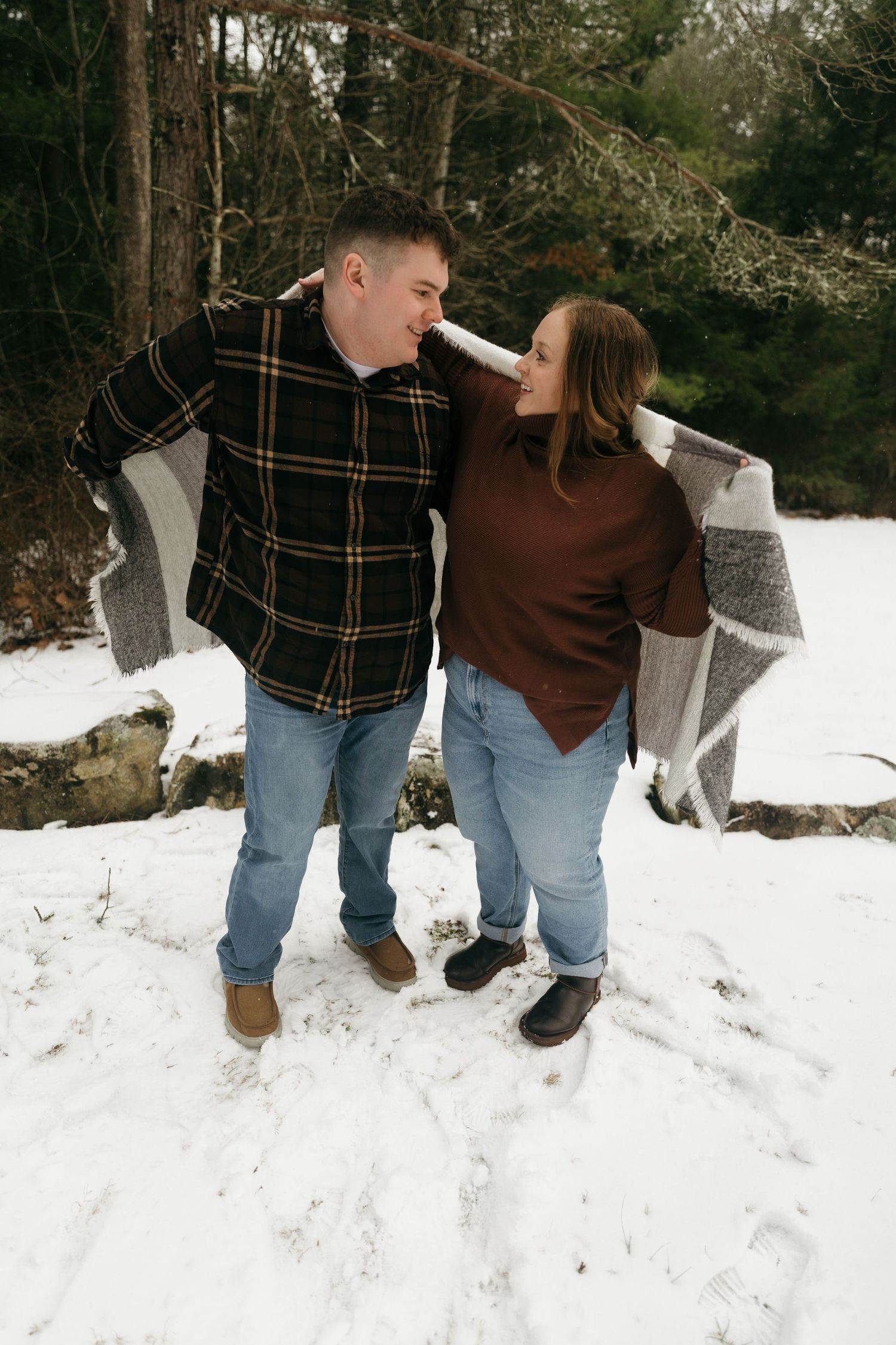 A couple wrapped in a grey blanket laughing together in a snowy forest.
