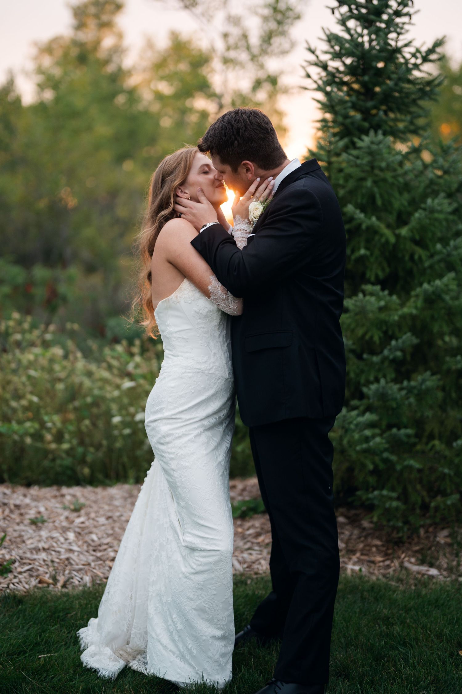 Bride and groom kissing at golden hour sunset in lush garden setting, bride in fitted lace gown.
