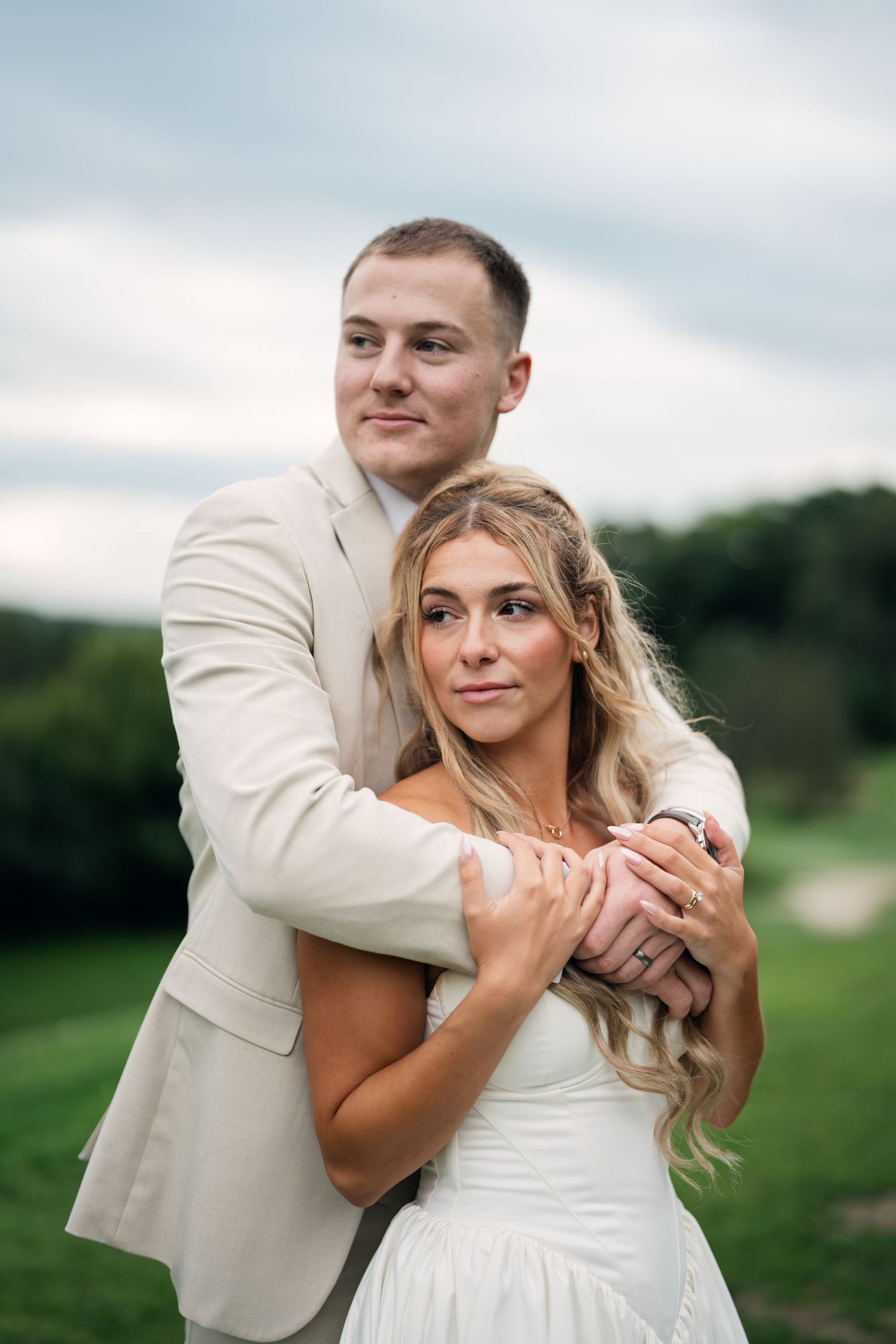 A groom in a beige suit embraces his bride in a white dress outdoors on their wedding day.