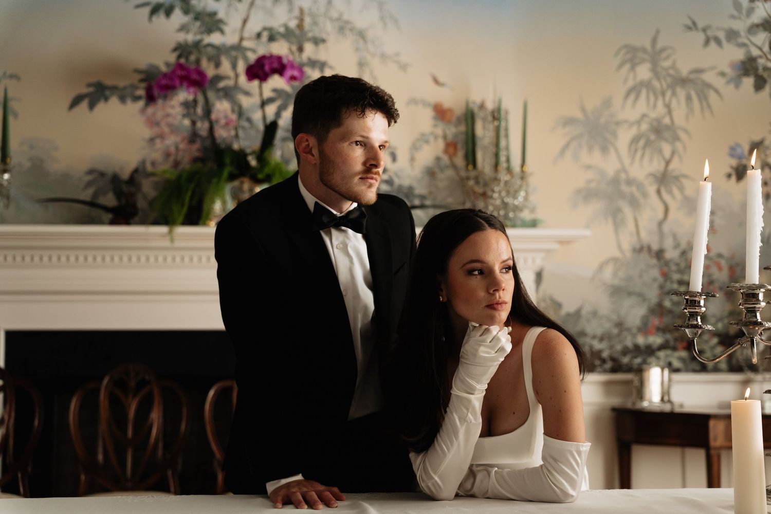 Elegant couple in formal attire pose together in a romantic indoor setting with candlelight and floral decor.