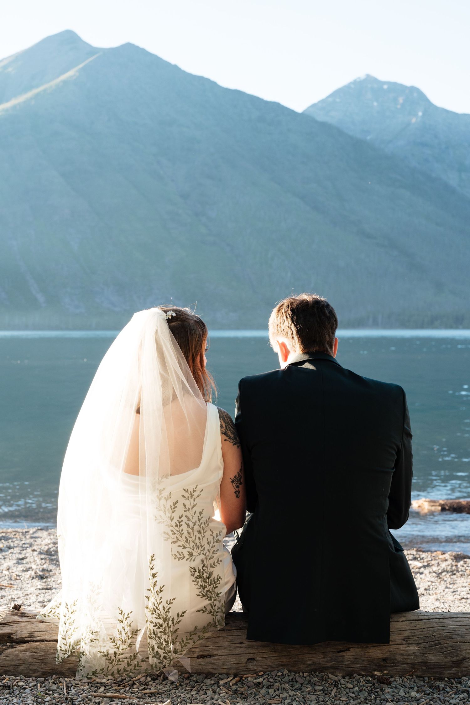 Bride and groom sitting together on a log by a mountain lake, viewed from behind, bride wearing veil with floral details.