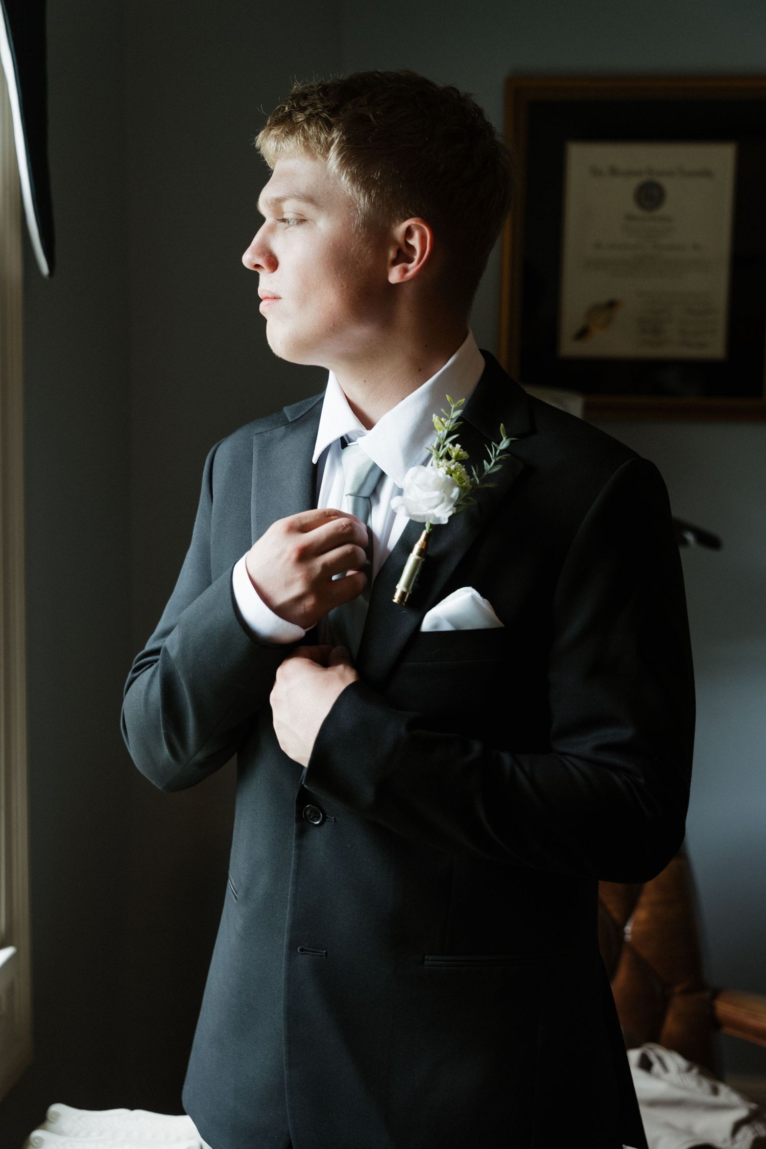 Young groom in dark suit adjusting boutonniere while gazing out window on wedding day.