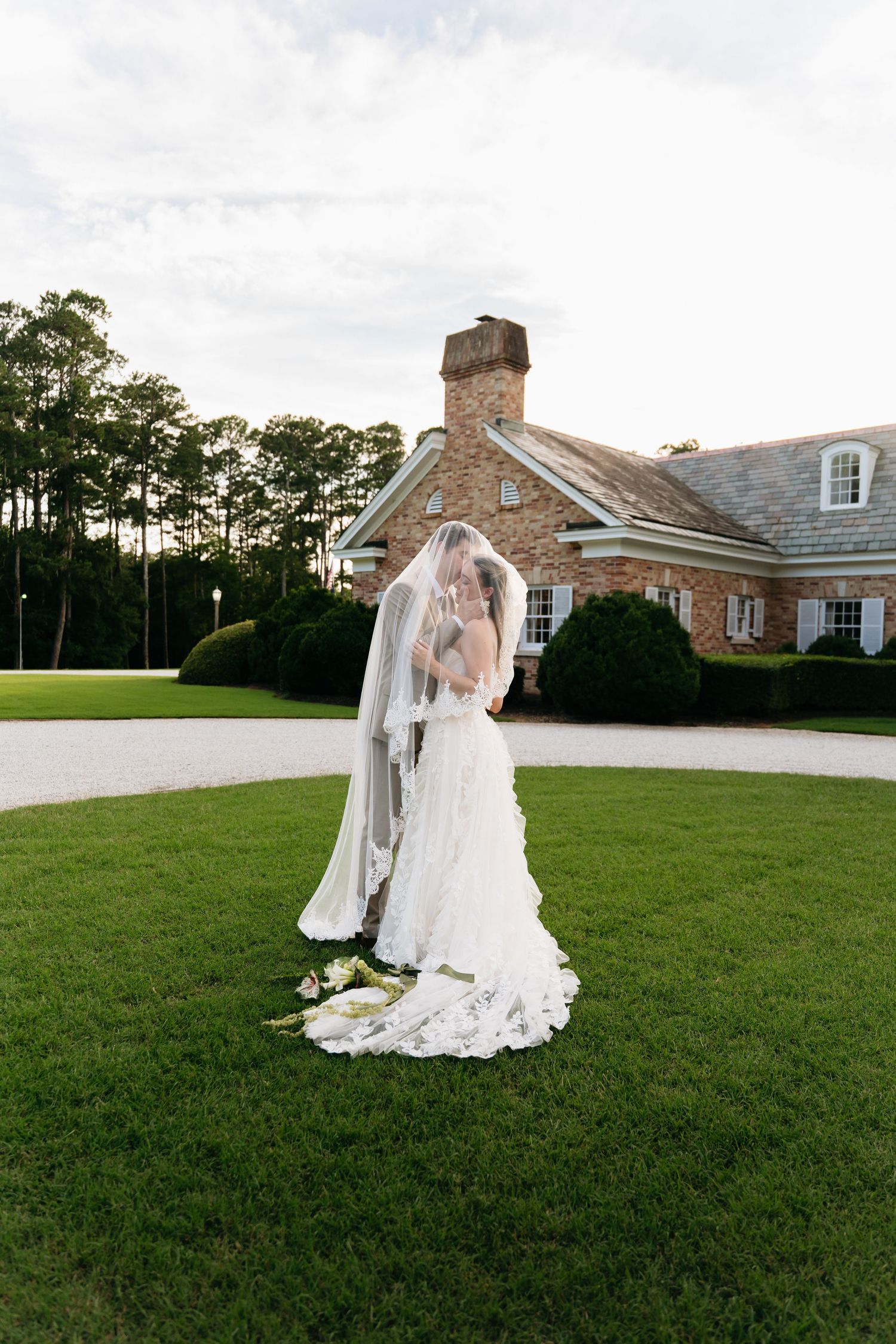 A bride in a flowing white gown and veil poses gracefully on a vast green lawn with a brick building in the background.