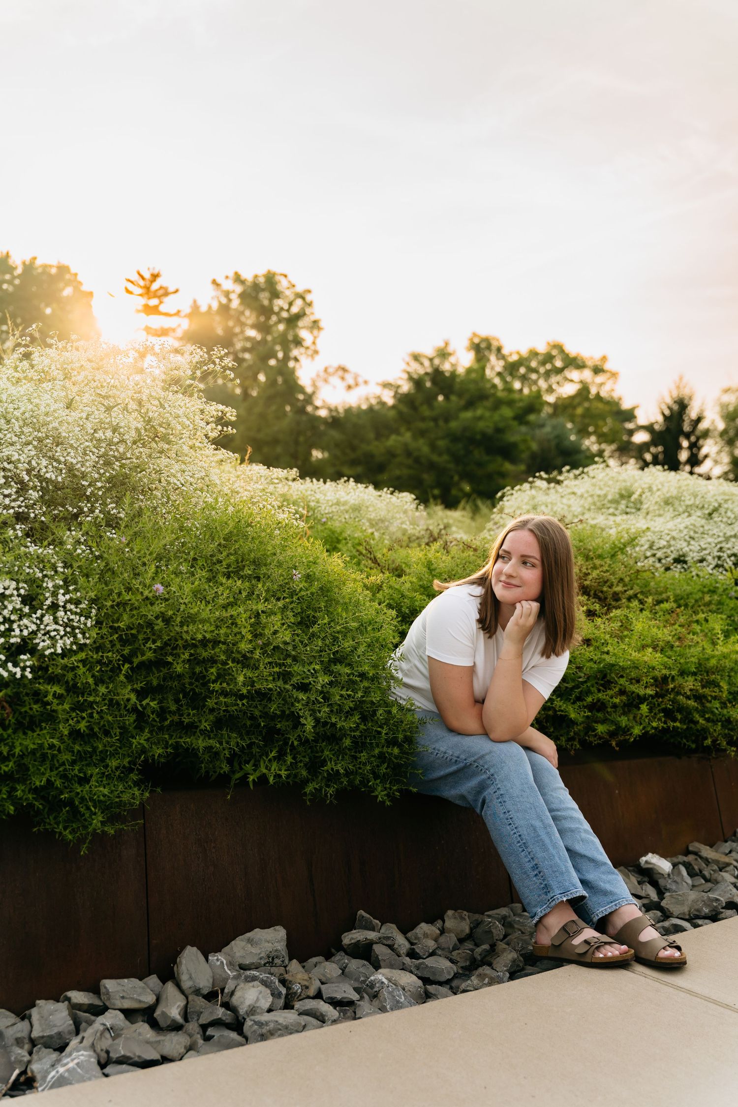 A person in casual attire sits on a stone wall surrounded by blooming white flowers during golden hour sunset.