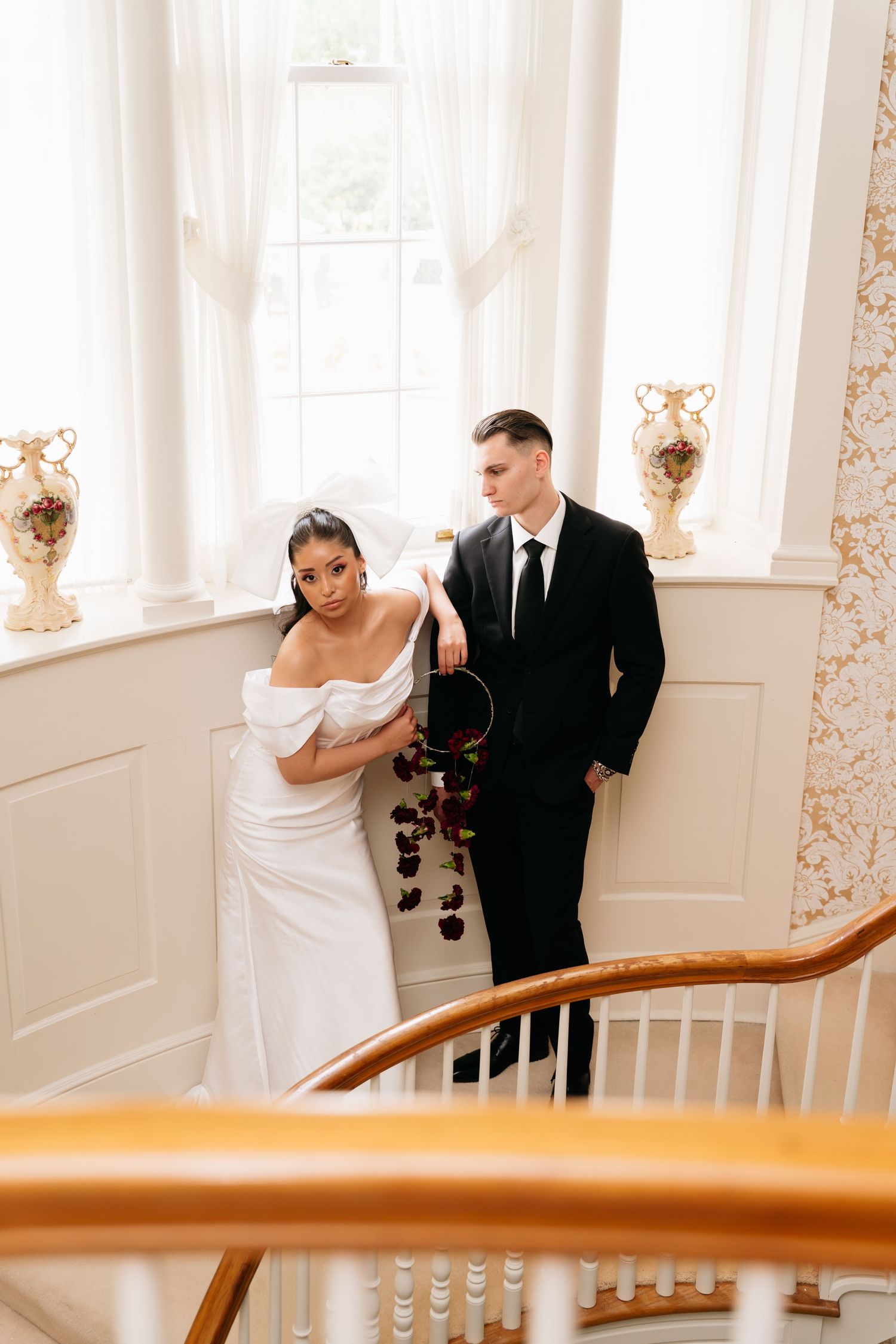 Bride in white off-shoulder gown and groom in black suit stand on elegant staircase by bright windows.