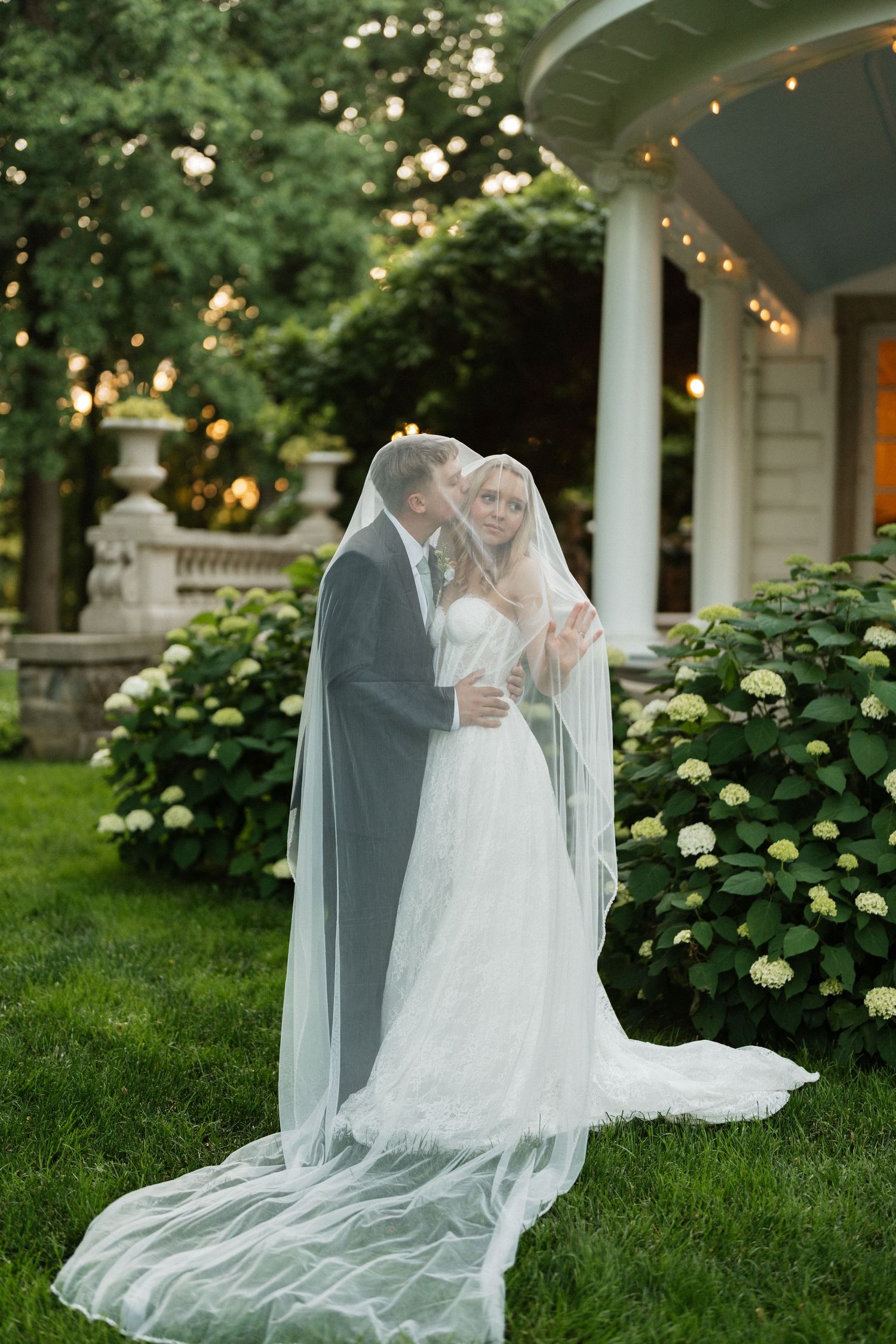 Bride and groom sharing a romantic kiss under a flowing veil in a lush garden with hydrangeas and string lights.