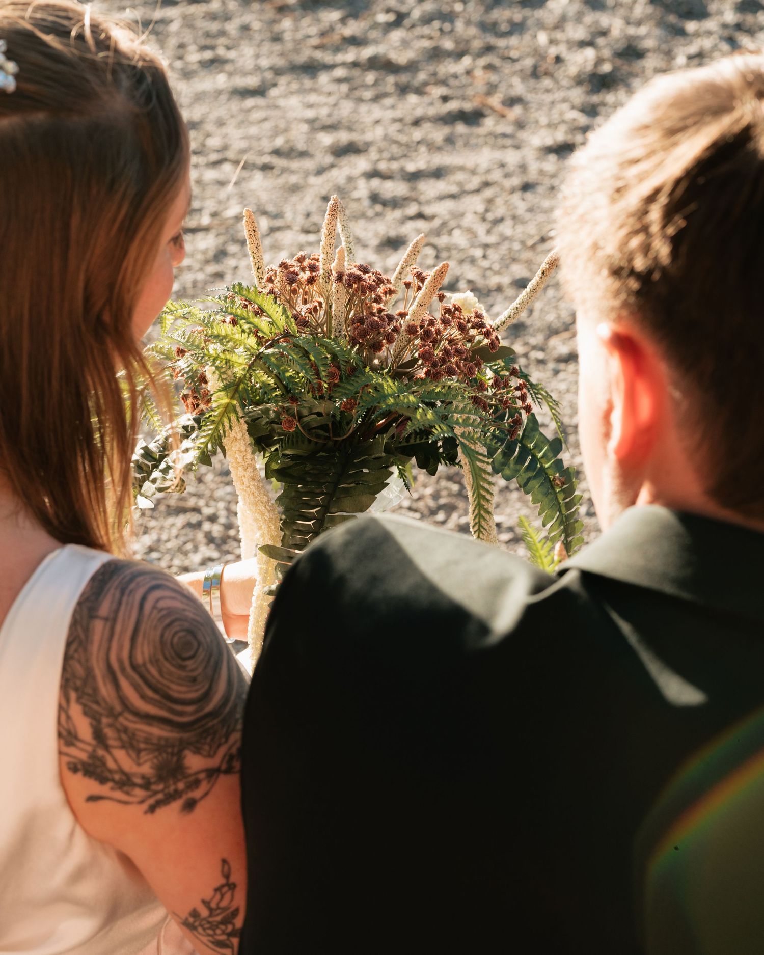 Bride with floral tattoo holding rustic wedding bouquet of ferns and berries beside groom in black suit.