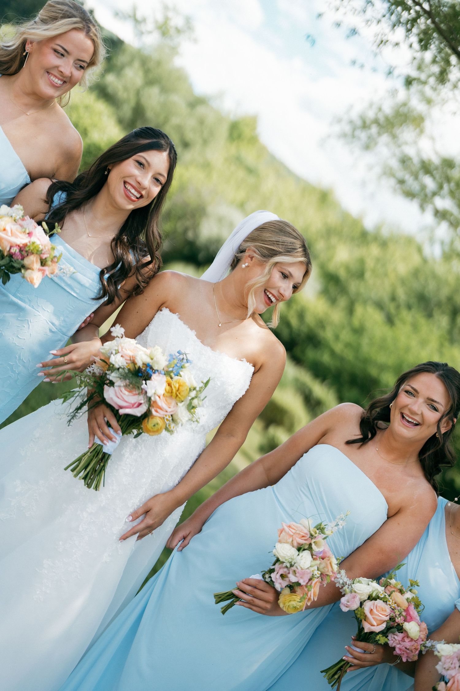 Bride in white gown with bridesmaids in light blue dresses holding colorful bouquets outdoors.