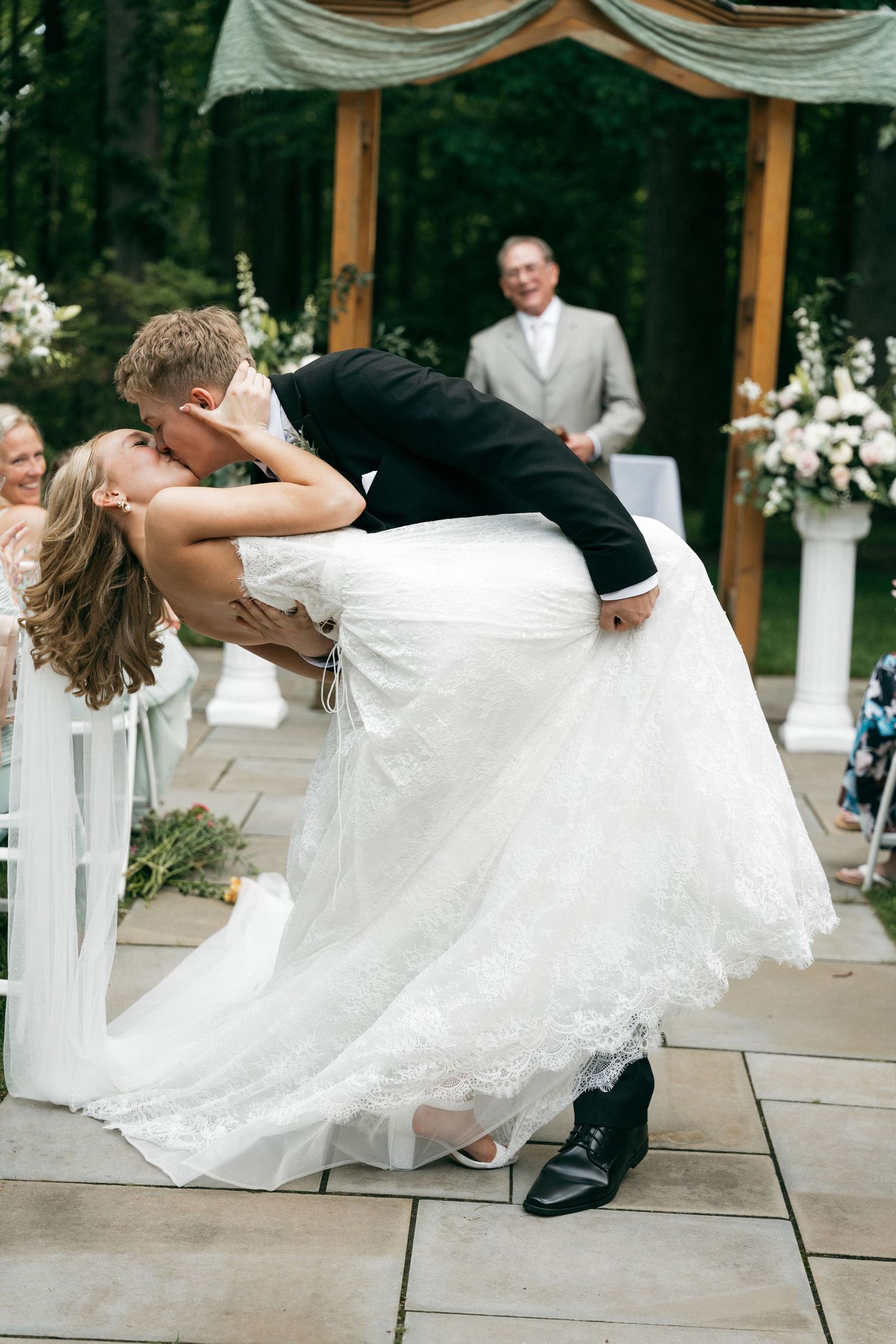 Groom dips and kisses bride in lace wedding gown at outdoor ceremony altar surrounded by floral arrangements.