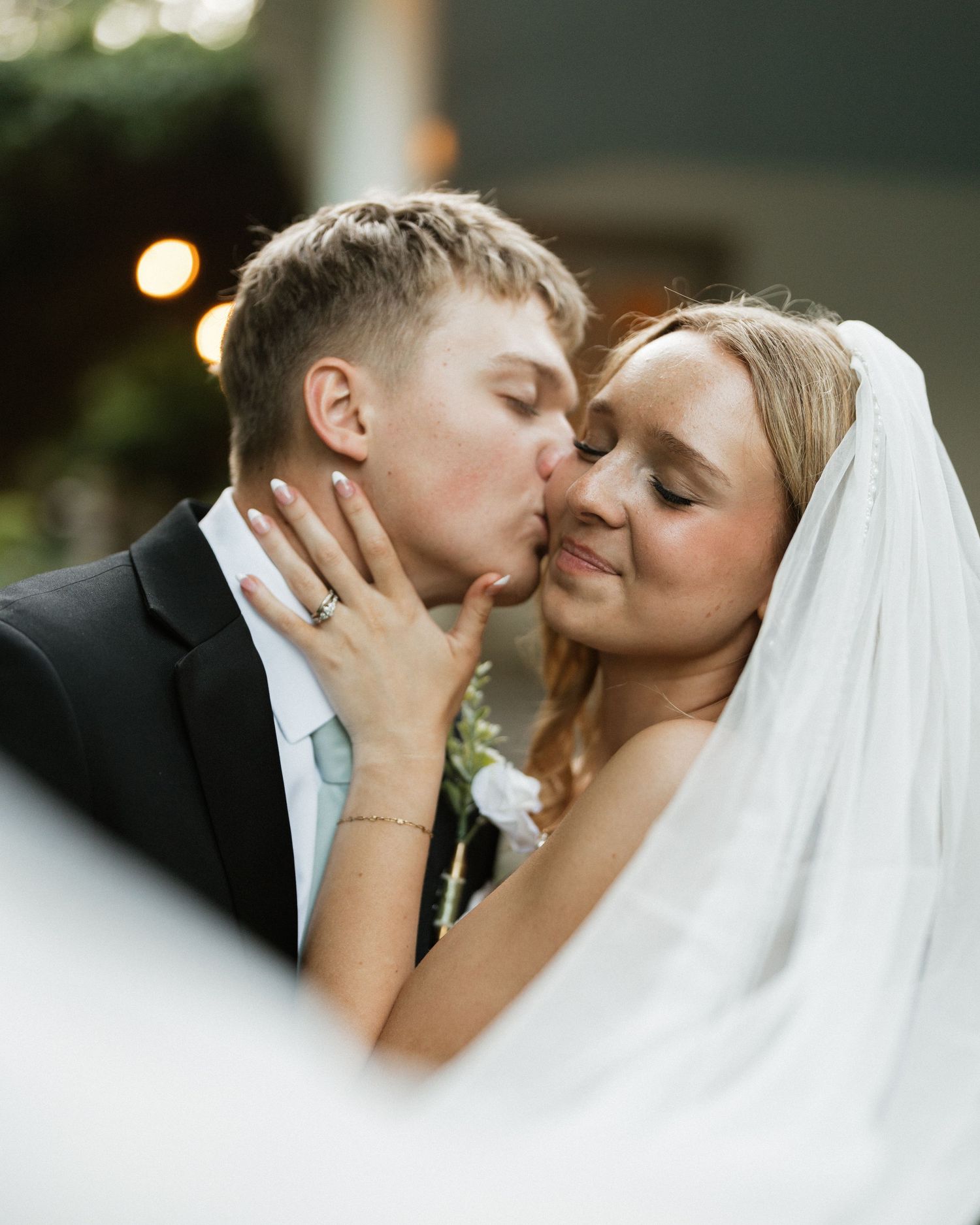 Groom tenderly kisses bride on cheek as she smiles softly during romantic outdoor wedding portrait.