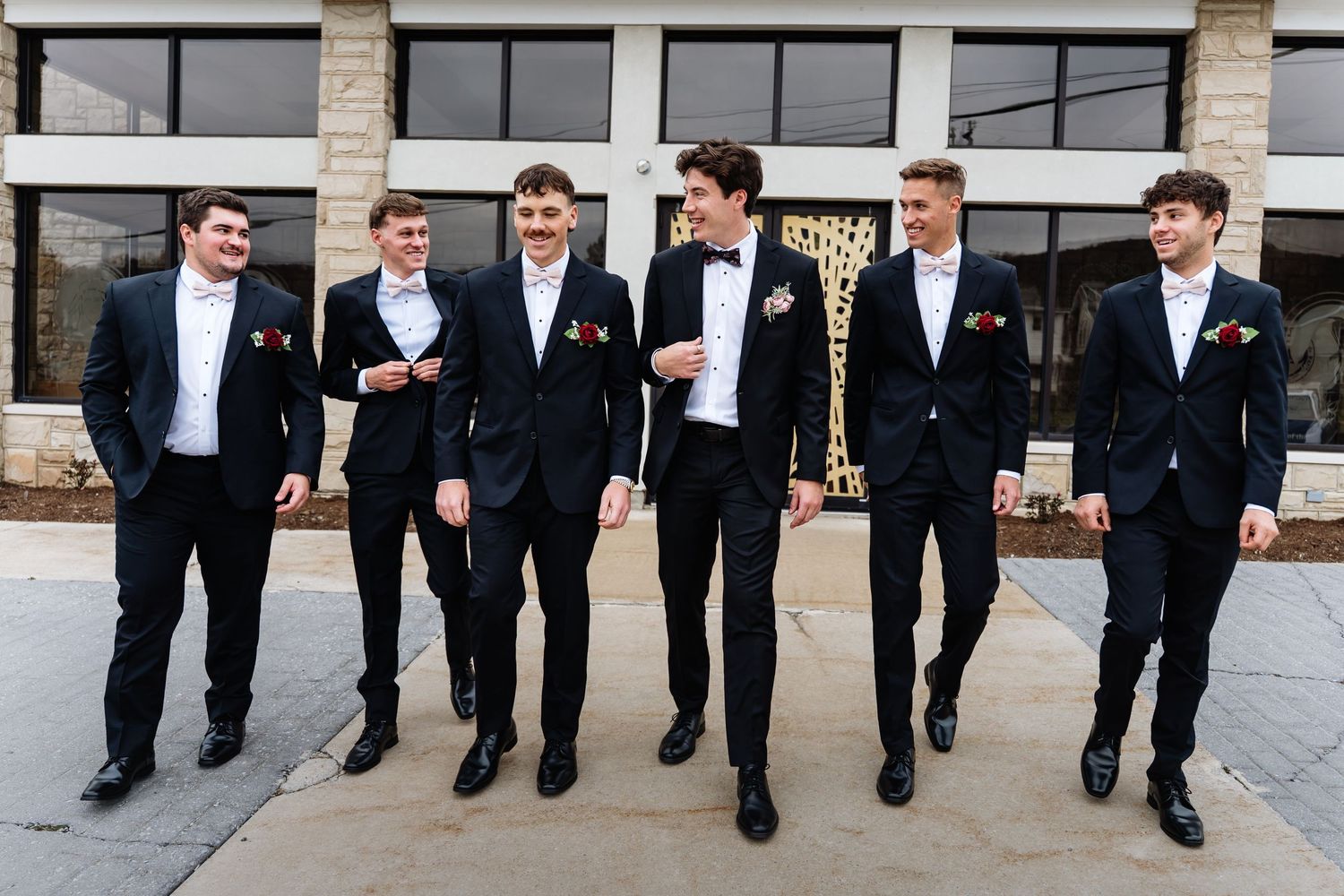 Groomsmen in black suits walk in a line on a stone walkway showing their colorful socks.