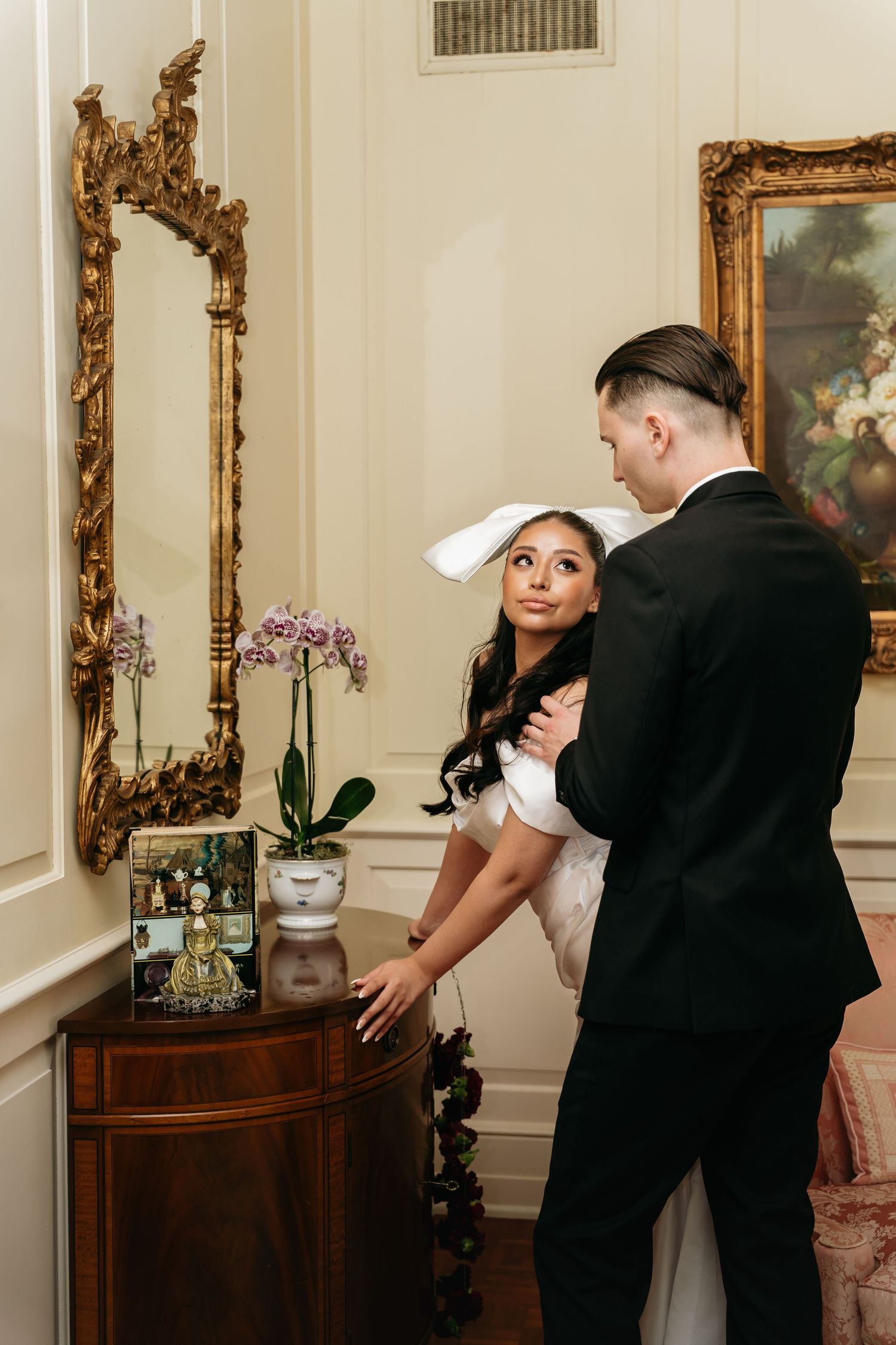 Bride in white gown and bow looks up at groom in black tuxedo near ornate gold mirror and antique wooden dresser.