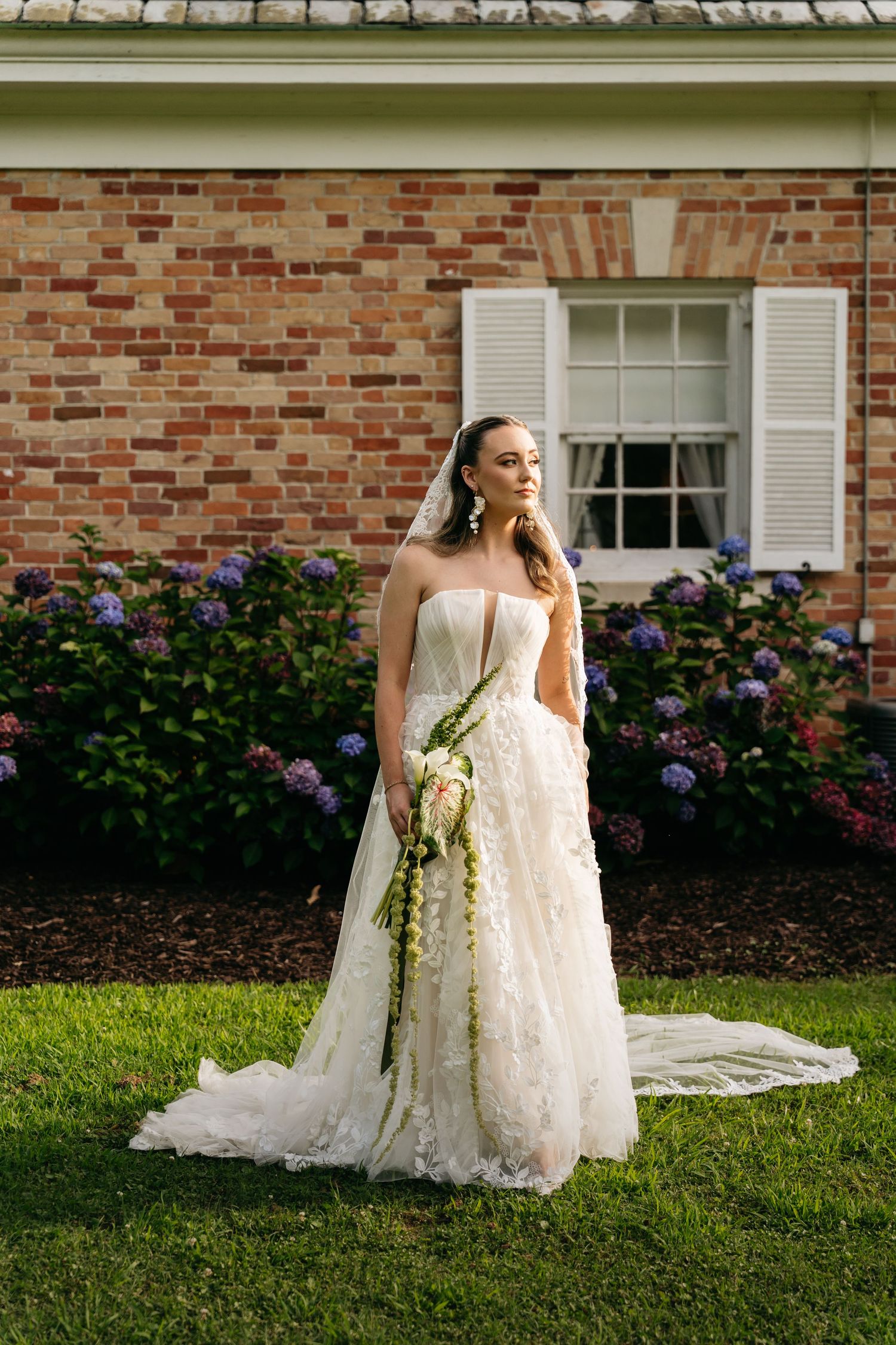 Bride in lace ball gown with veil holds cascading bouquet before brick wall with hydrangeas.