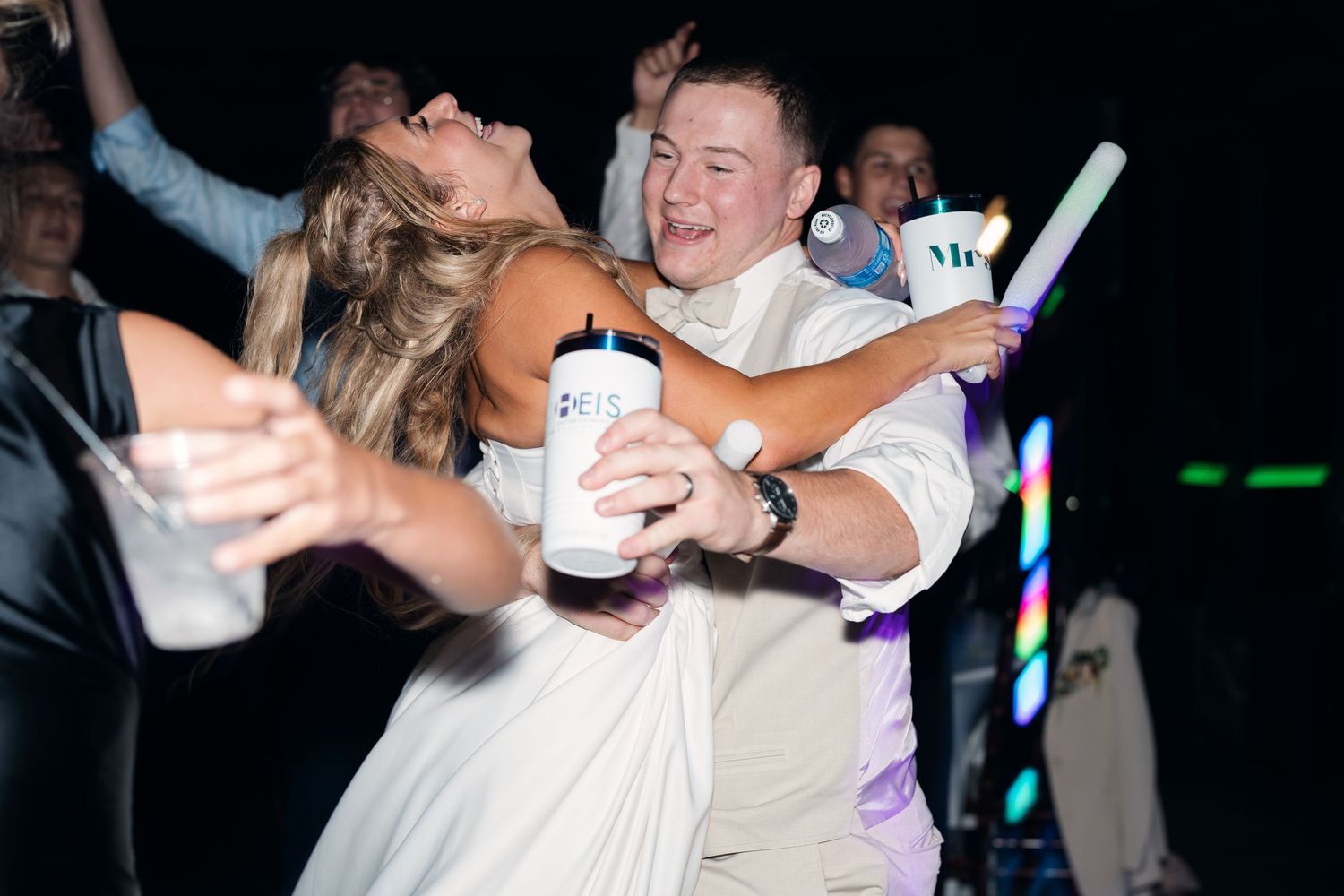 Bride and groom dancing joyfully at wedding reception, holding light-up accessories on the dance floor.