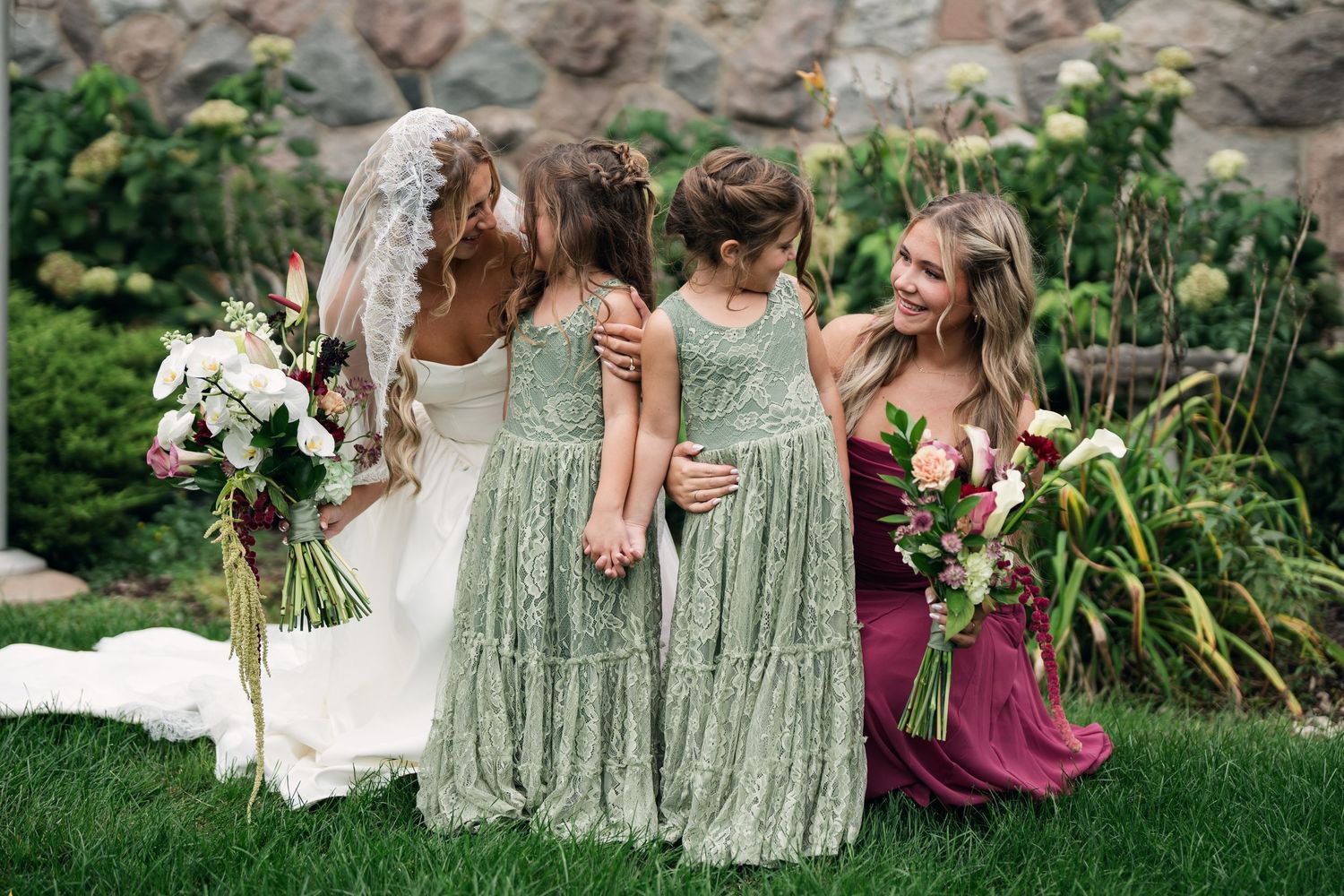 Bride in white gown with bridesmaids in sage green and burgundy dresses holding floral bouquets in a garden setting.