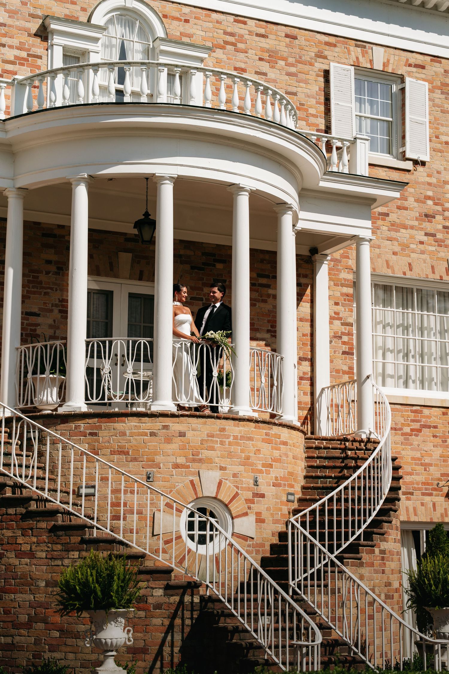 Elegant curved white columns and wrought iron railings adorn the front porch of a classic brick home.