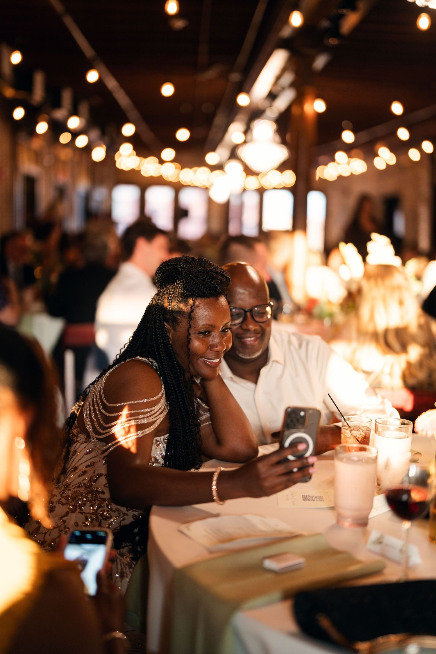 Couple smiling at phone during wedding reception with warm string lights glowing in elegant venue.