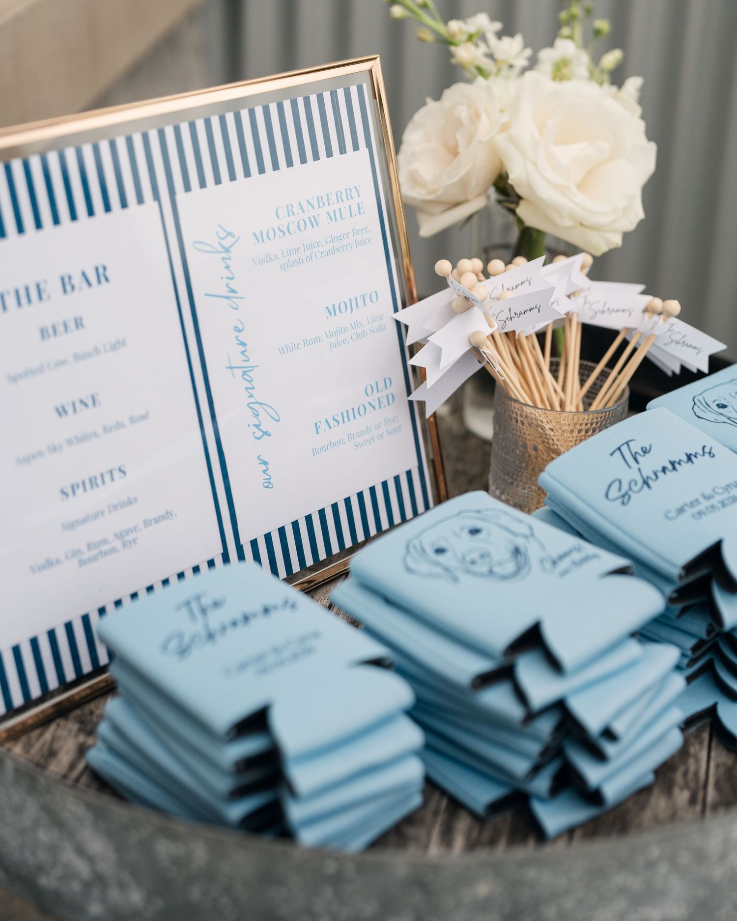 Blue wedding bar setup with striped menu sign, custom napkins, and floral arrangements for an elegant event.