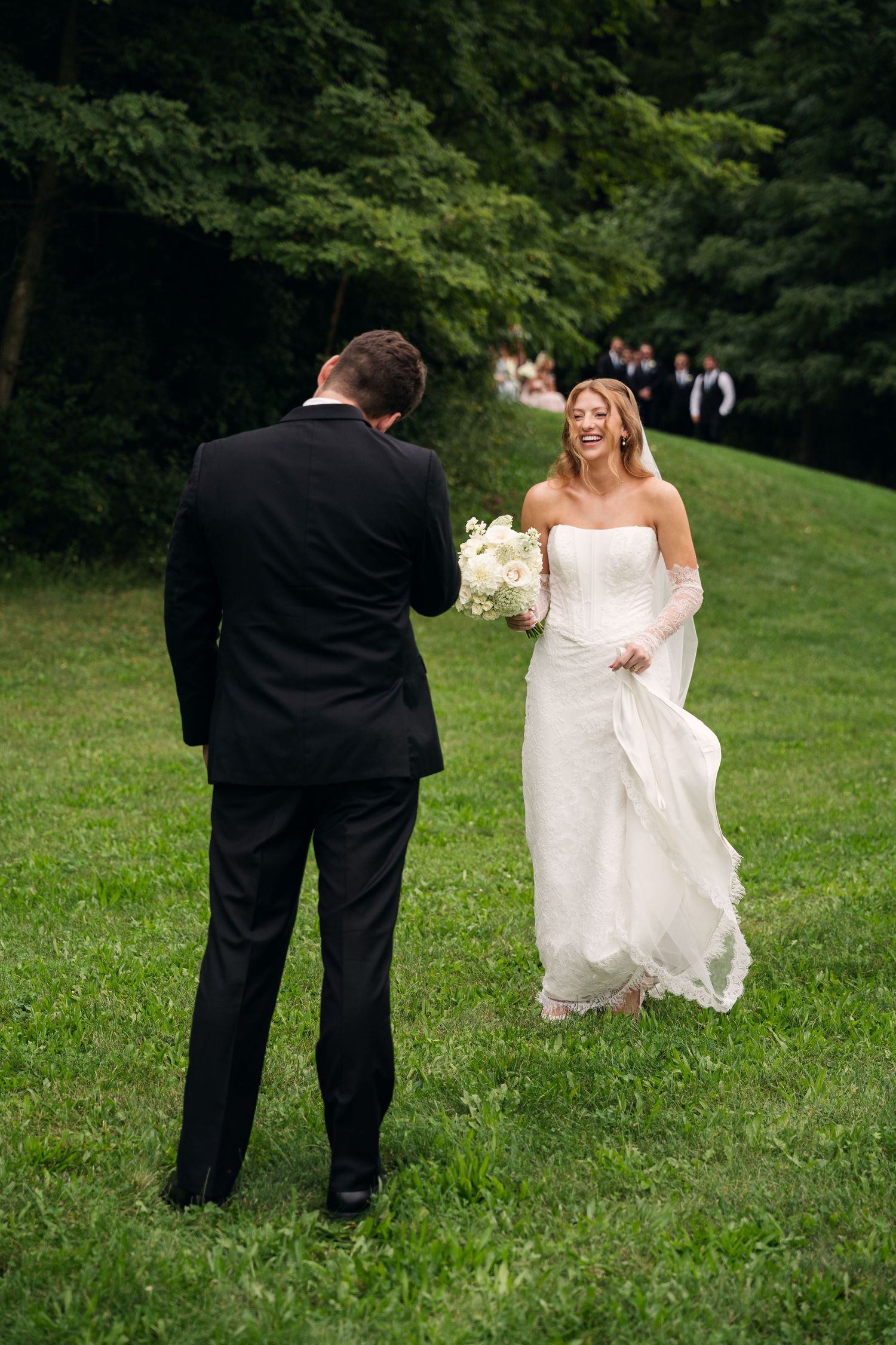 Smiling bride in strapless white gown holding bouquet approaching groom on a lush green lawn.