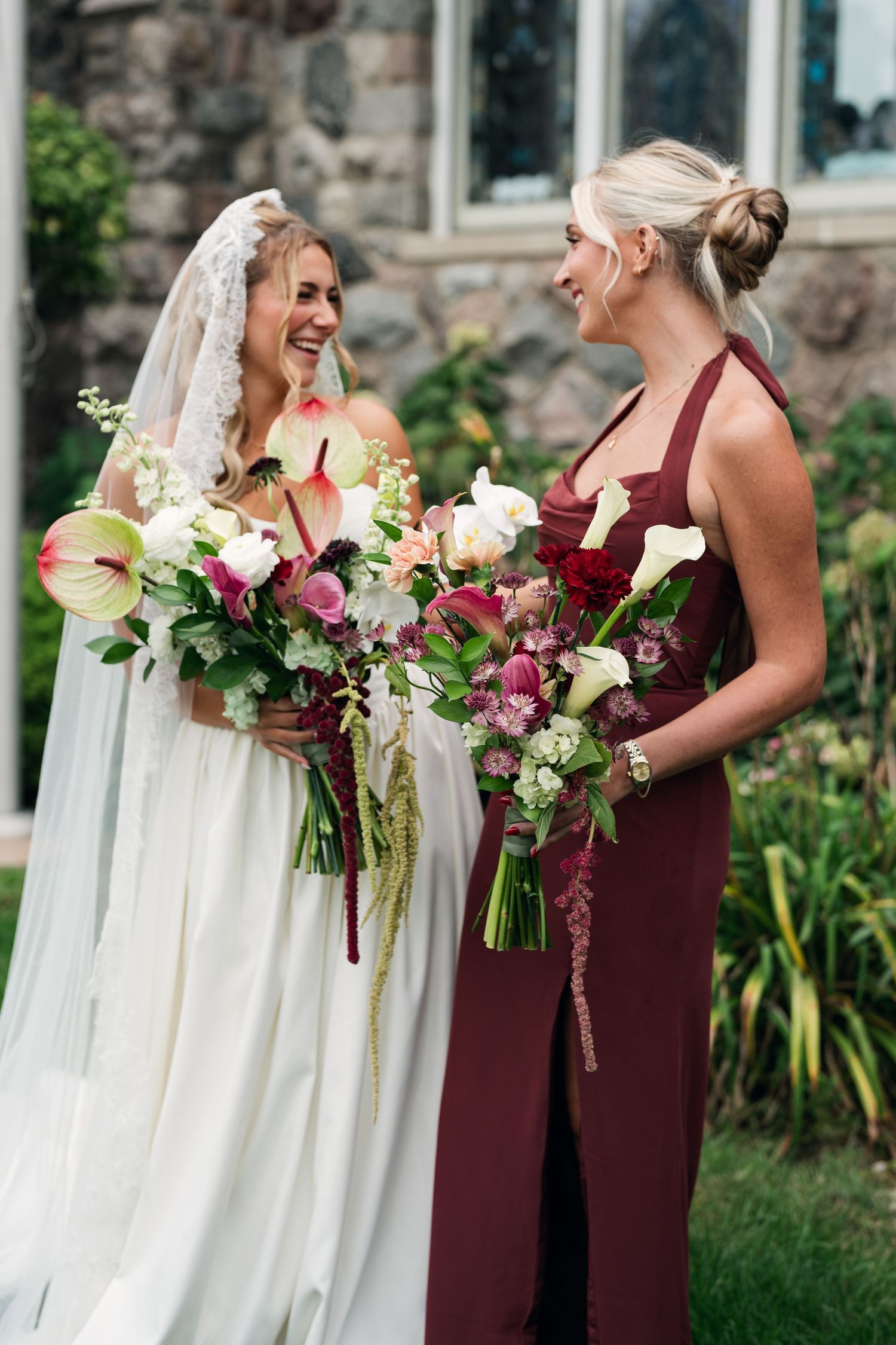Bride in white gown and veil laughs with bridesmaid in burgundy dress, both holding lush floral bouquets outdoors.
