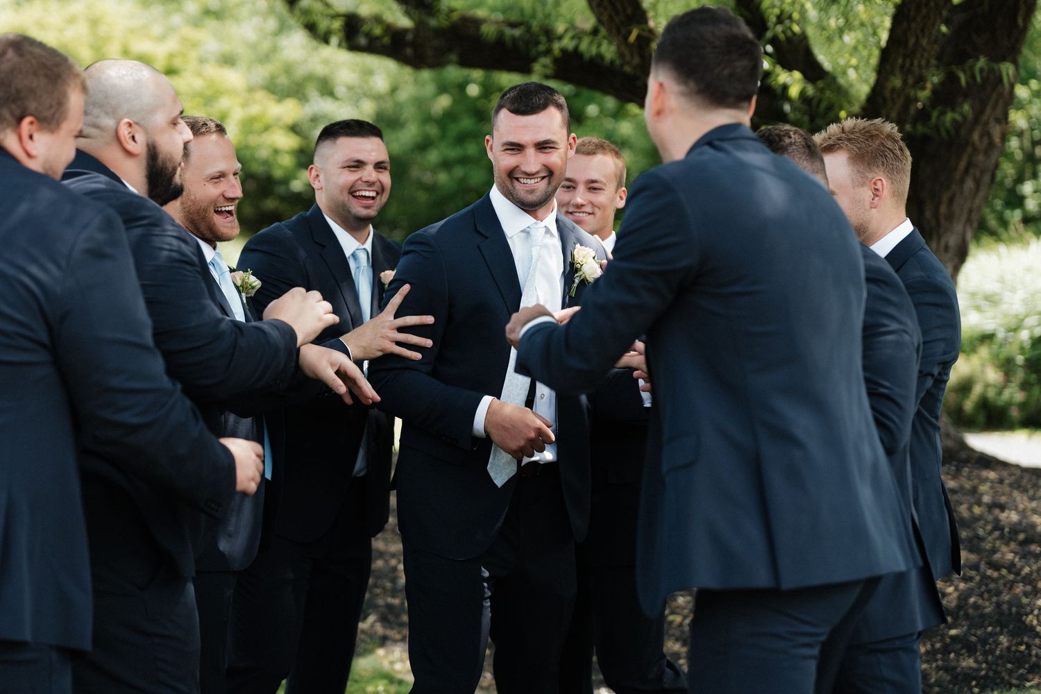 Groomsmen in navy suits laughing and celebrating with the groom outdoors at a wedding.