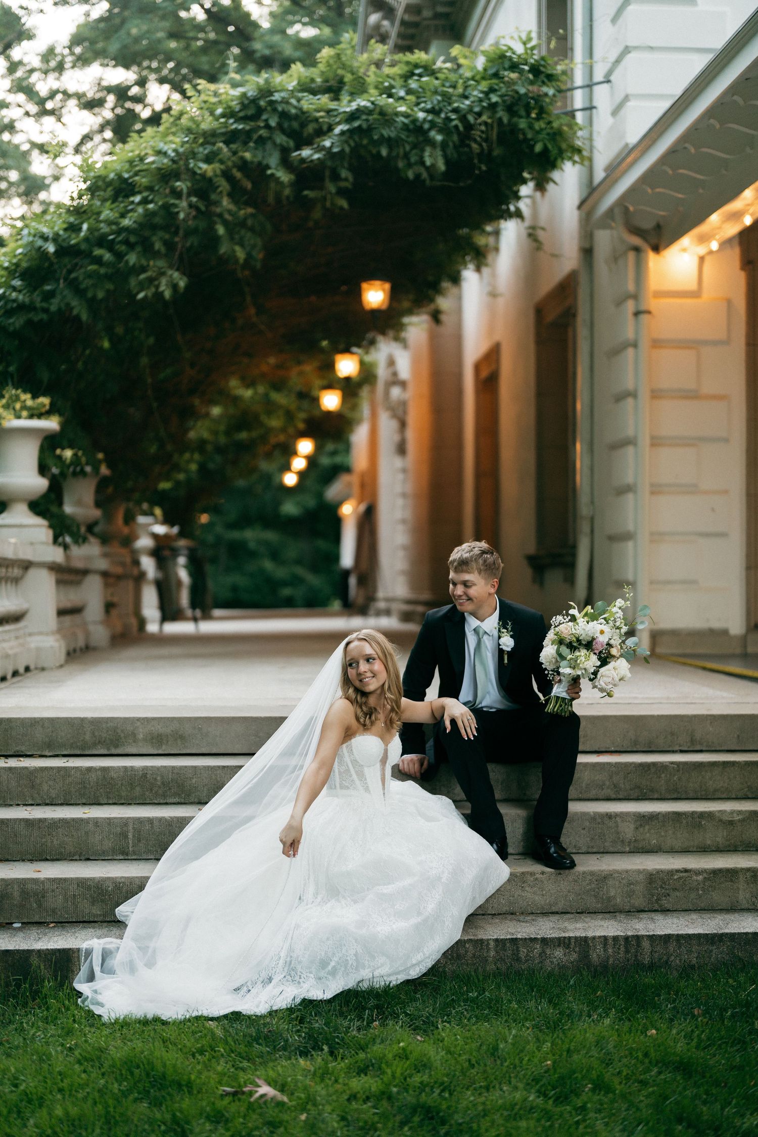 Bride in flowing white lace gown and groom with bouquet sit on stone steps of elegant venue at dusk.