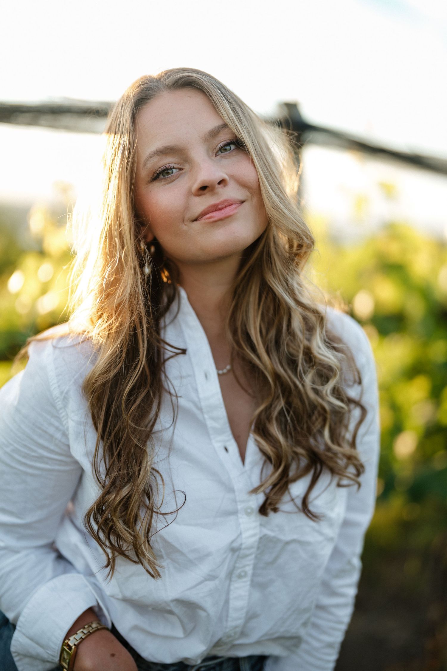 Young woman with long wavy blonde hair smiling outdoors in white shirt with green foliage background.