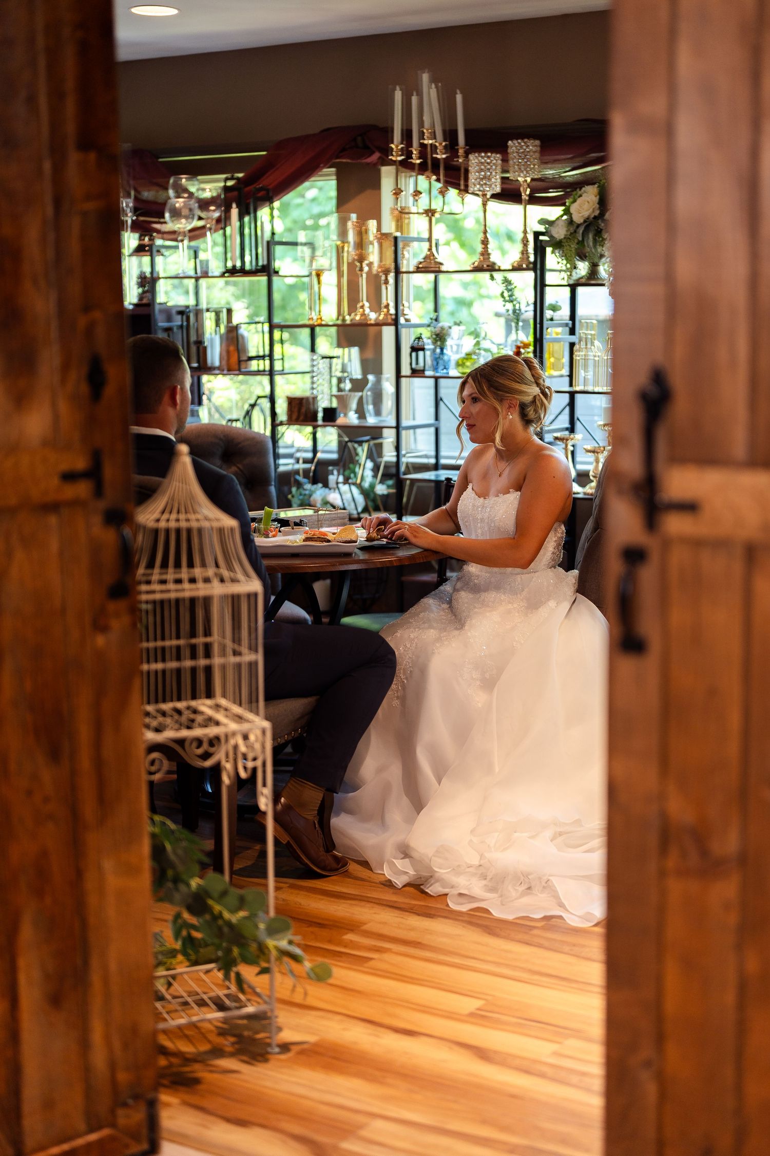 Bride and groom sit together at a decorated table in a rustic venue with wood floors and elegant chandelier lighting.