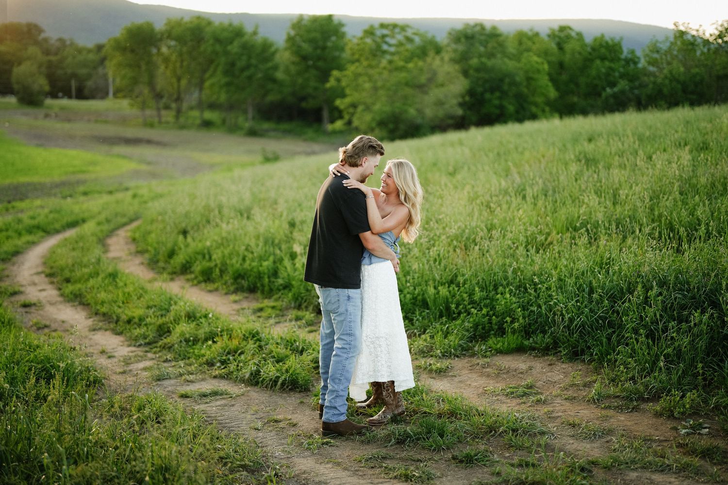 Couple embraces romantically on a winding dirt path surrounded by lush green fields at sunset.