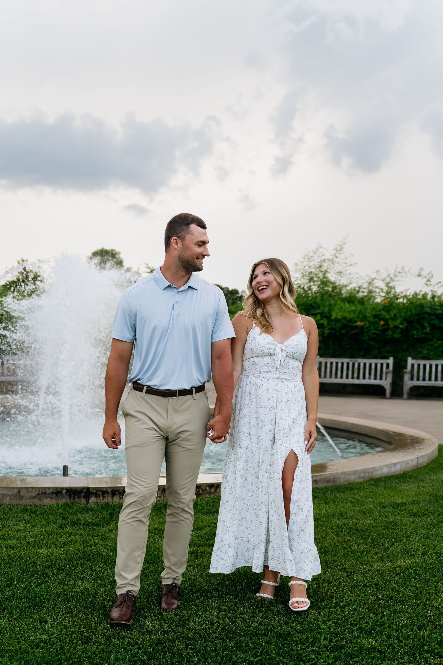 A couple in light summer attire walks together on a grassy lawn near a decorative fountain.