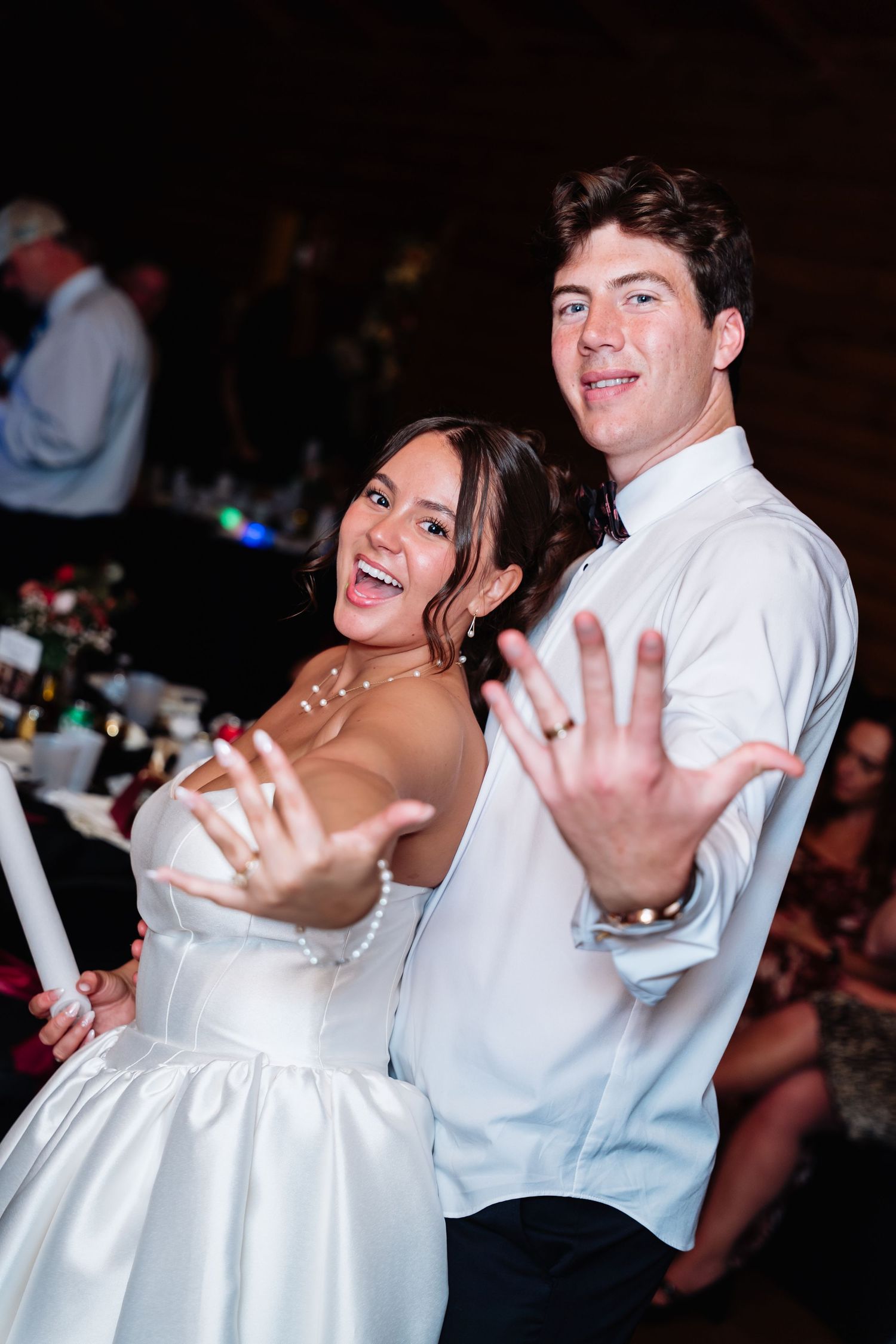 Newlyweds joyfully show off their wedding rings at their reception, bride in white gown, groom in white dress shirt.