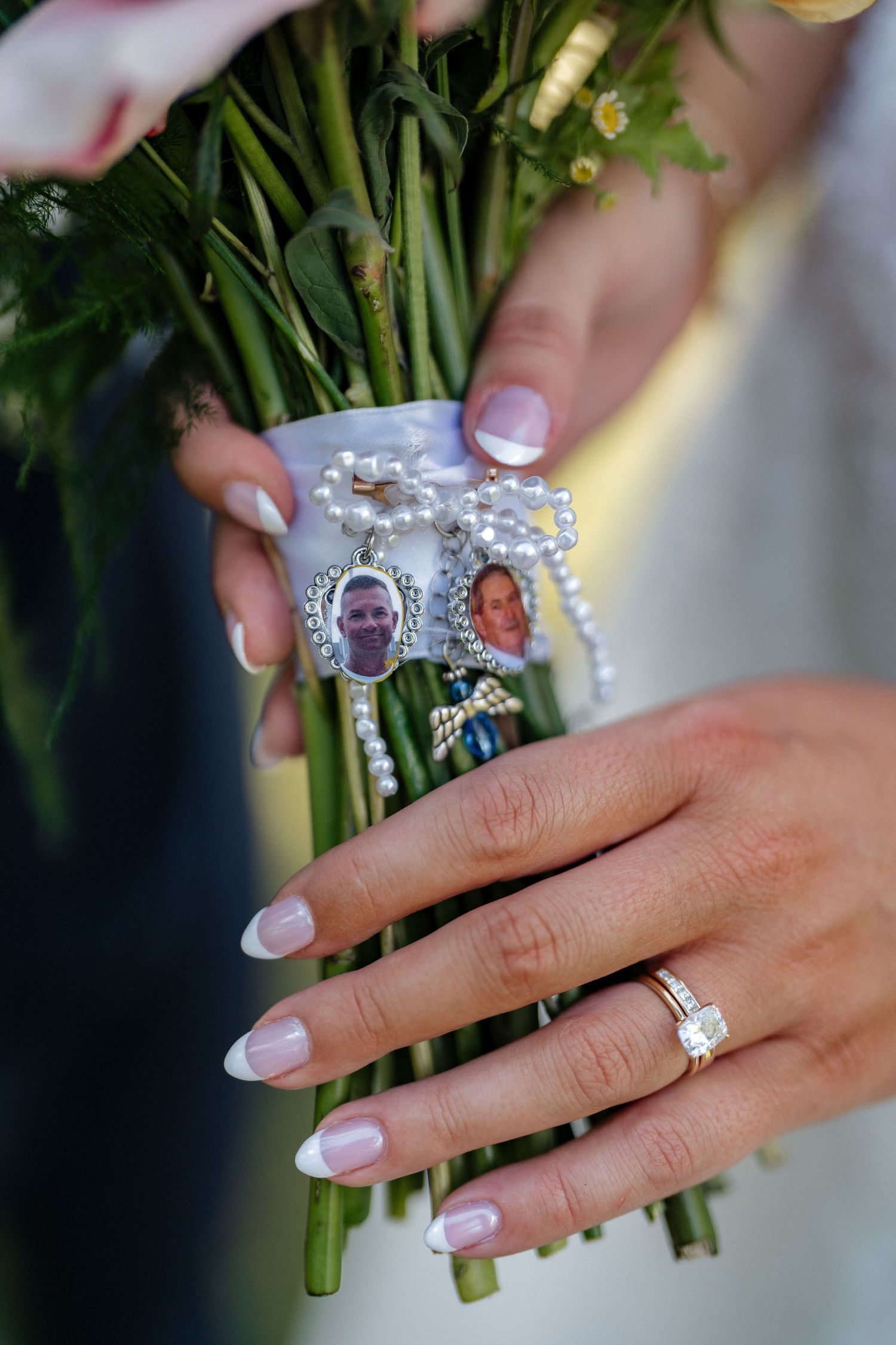 Bride holding wedding bouquet with memorial photo charms and pearl rosary, wearing diamond engagement ring with French manicure.