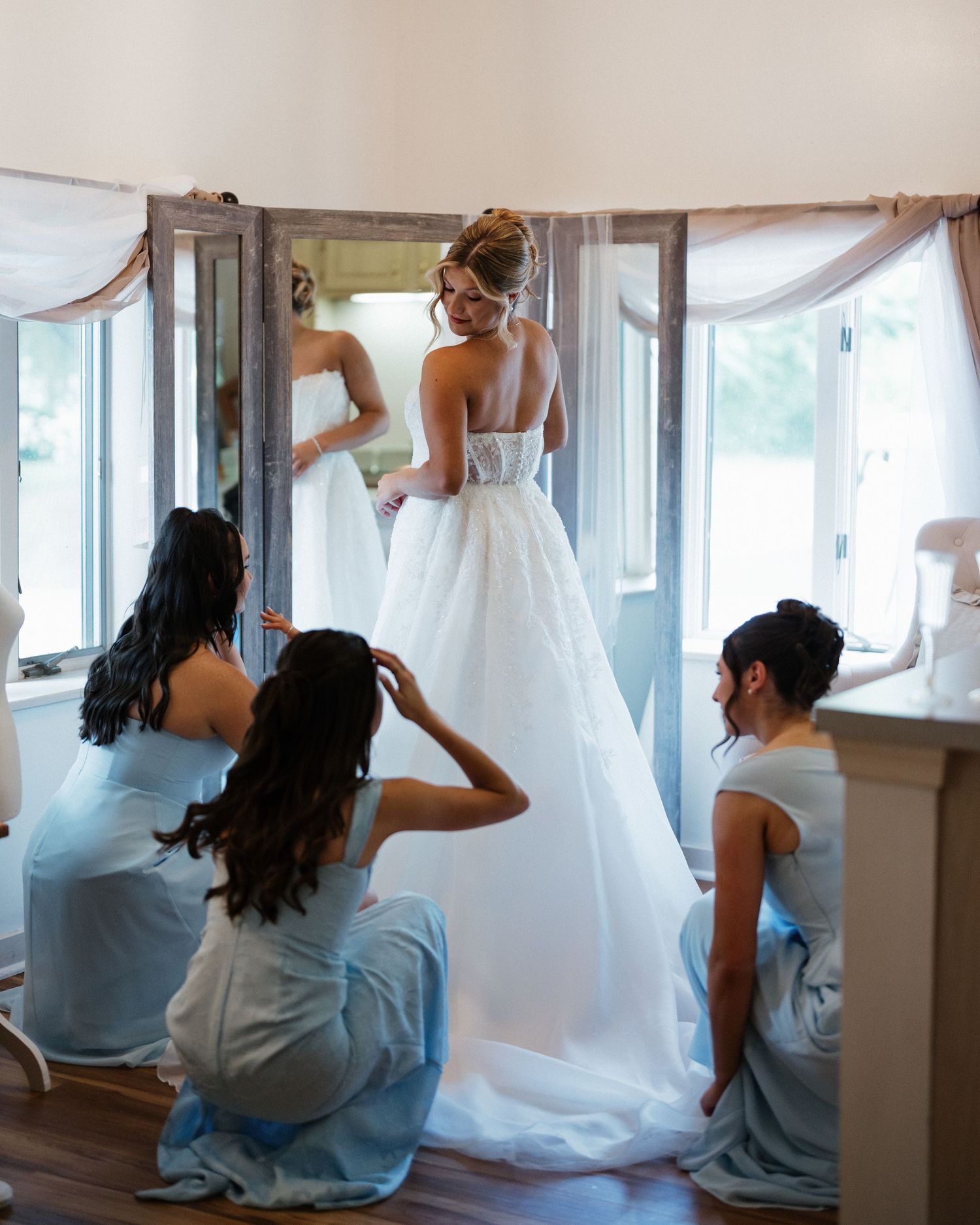 Bride in white gown being helped by bridesmaids in blue dresses near elegant draped windows before wedding.