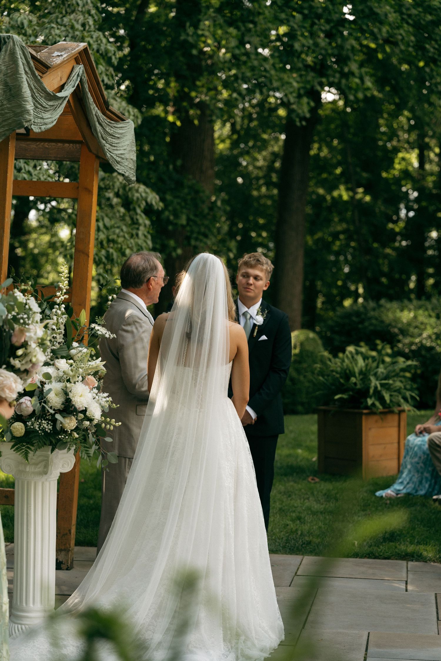 Bride in white gown and veil stands at outdoor altar facing groom in black suit during garden wedding ceremony.